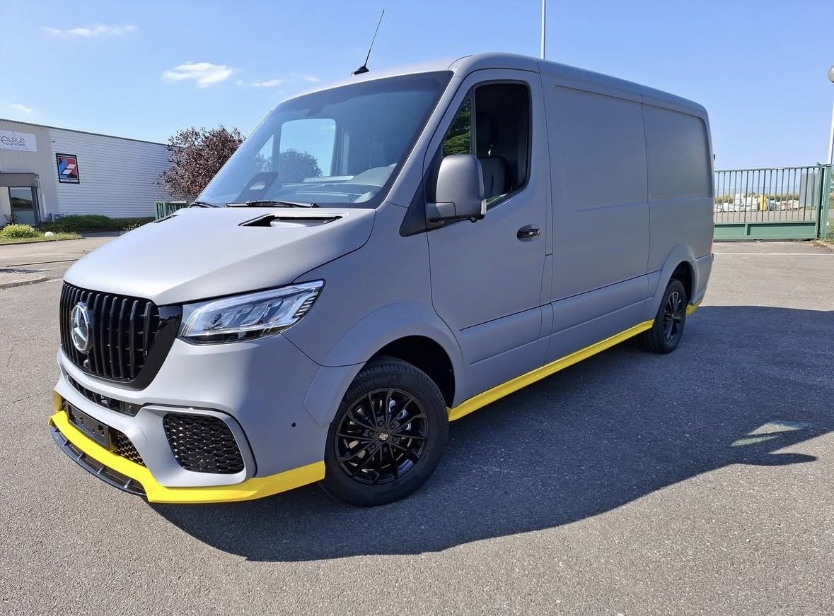 Véhicule utilitaire gris avec des accents jaunes, stationné sur une surface asphaltée, devant un bâtiment industriel.