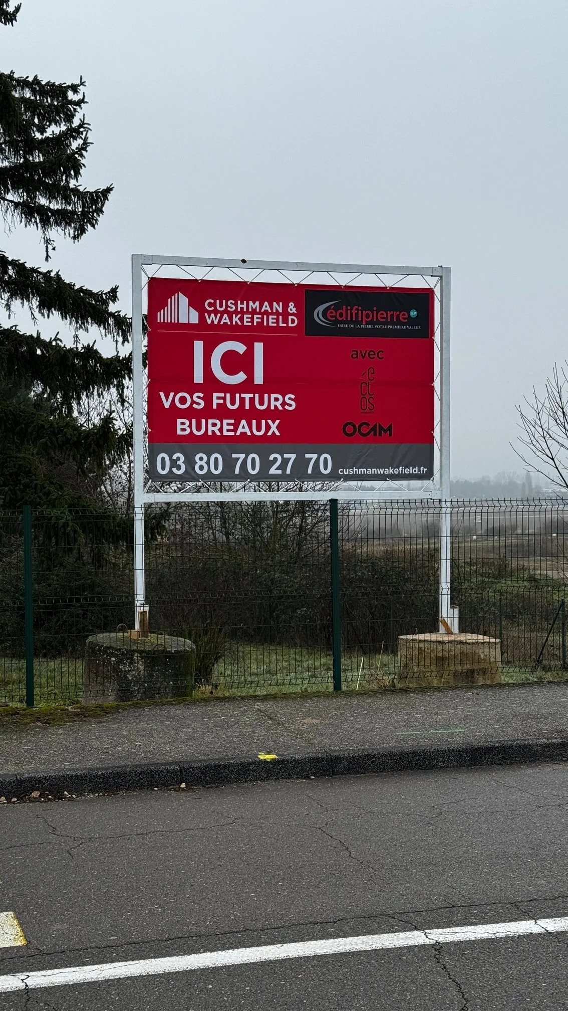 Panneau publicitaire rouge et noir pour Cushman & Wakefield, avec un message indiquant 'ICI VOS FUTURS BUREAUX', un numéro de téléphone, un site web et plusieurs logos.