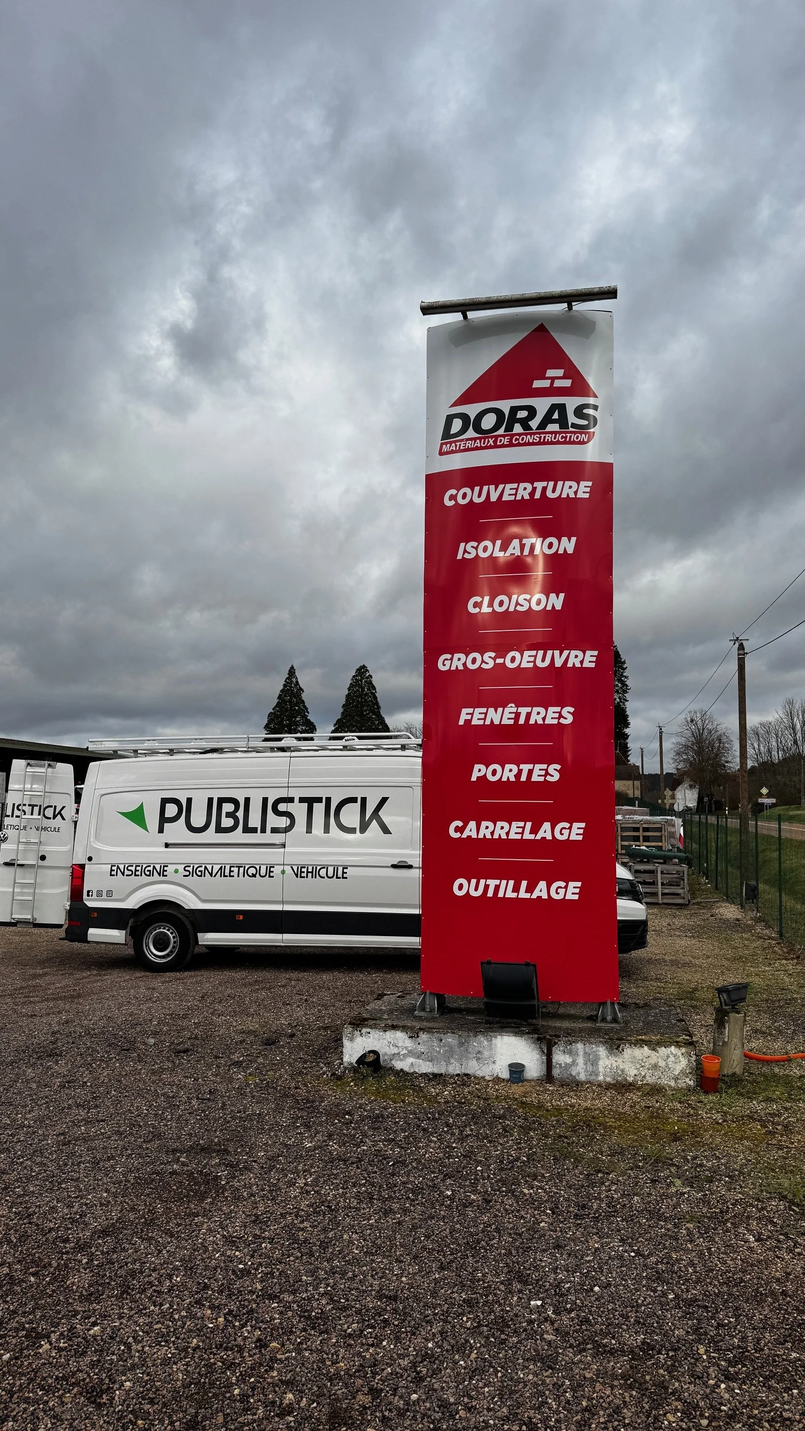 Panneau publicitaire rouge avec texte blanc listant des services de construction, tels que couverture, isolation, cloison, gros-oeuvre, fenêtres, portes, carrelage, outillage, situé à côté d'un camion blanc à l'apparence d'une camionnette.
