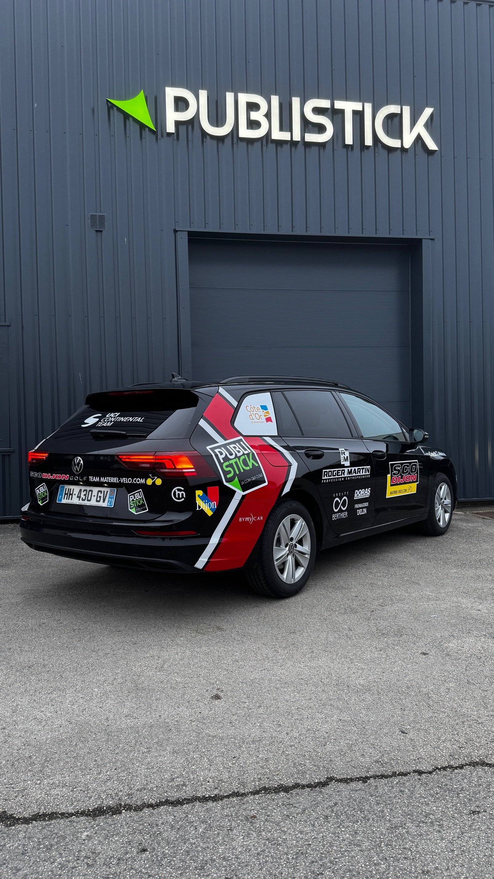 Voiture de course Volkswagen Polo avec des logos publicitaires, stationnée devant un bâtiment avec un panneau 'PUBLISTICK'.