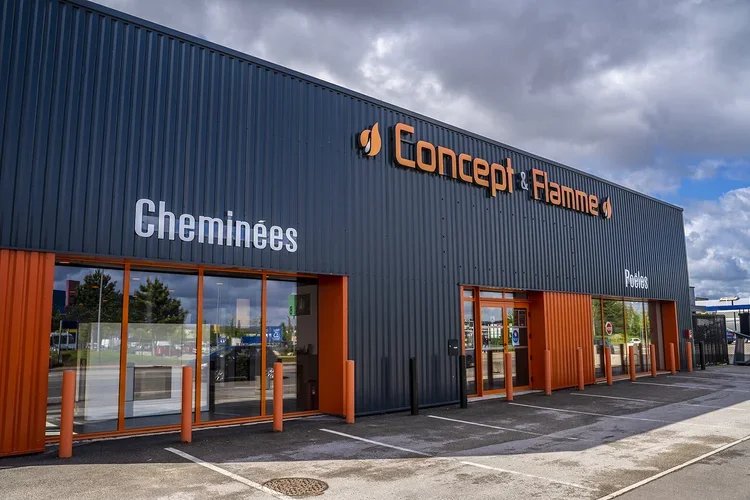 Un bâtiment commercial avec le nom "Concept & Flamme" en lettres orange et noires. Sur le bâtiment, on voit deux inscriptions : "Cheminées" à gauche et "Poêles" à droite. Il y a des fenêtres en verre et une petite zone de stationnement devant.