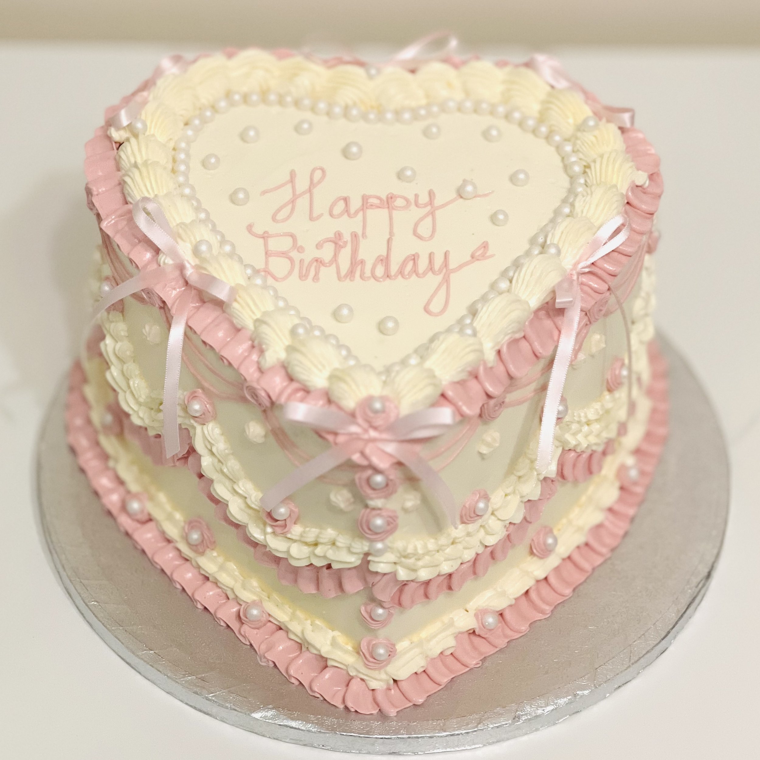 Vintage-style heart-shaped birthday cake in blush pink and ivory, decorated with pearls and ribbons
