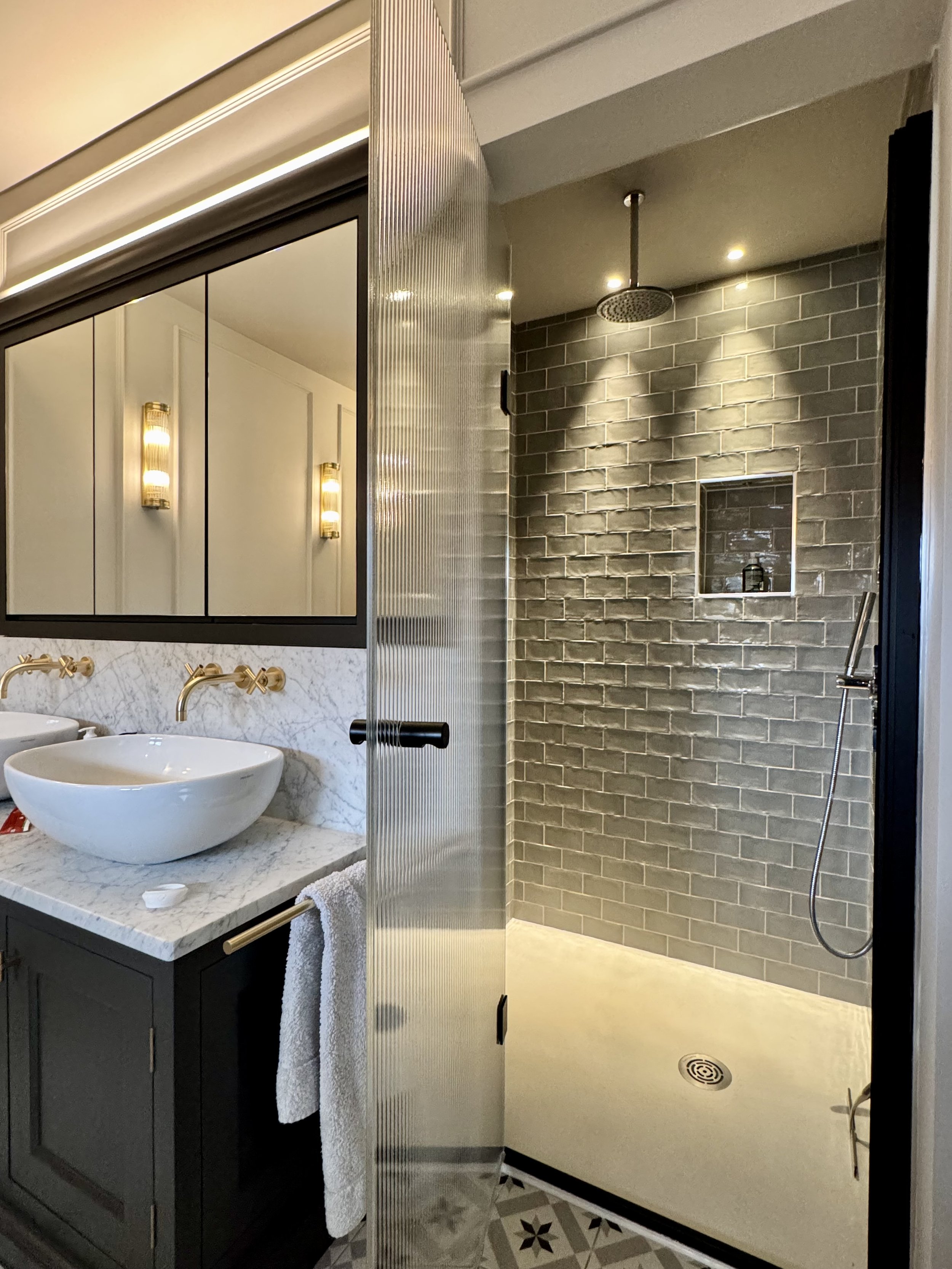 Modern bathroom with vanity and vessel sink, gold fixtures, lit wall sconces, and a walk-in shower with gray subway tile and a rainfall showerhead.