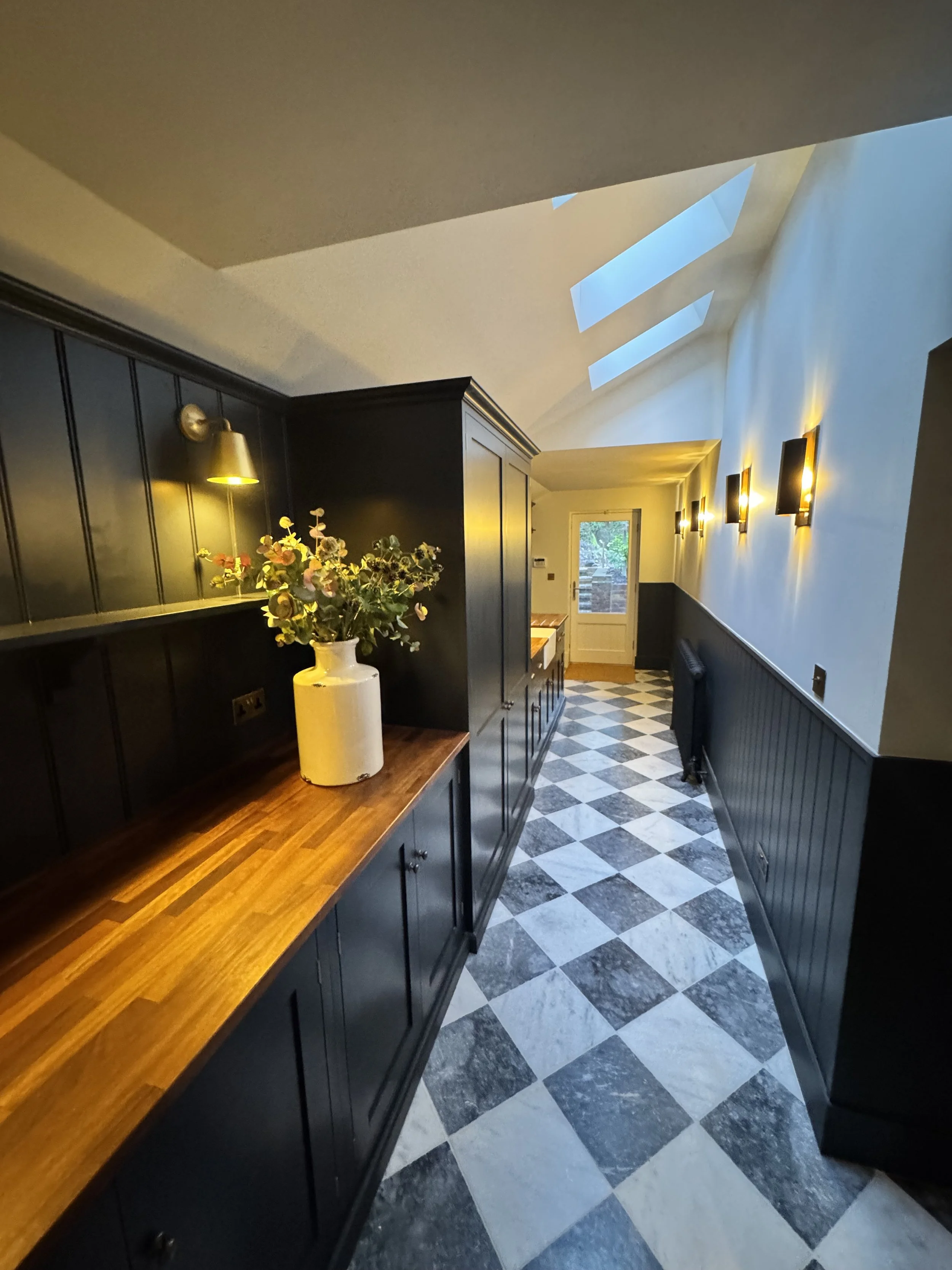 A kitchen corridor with black cabinetry, a wooden countertop, and a checkered black and white tile floor. The space has wall-mounted lights and skylights providing natural light.