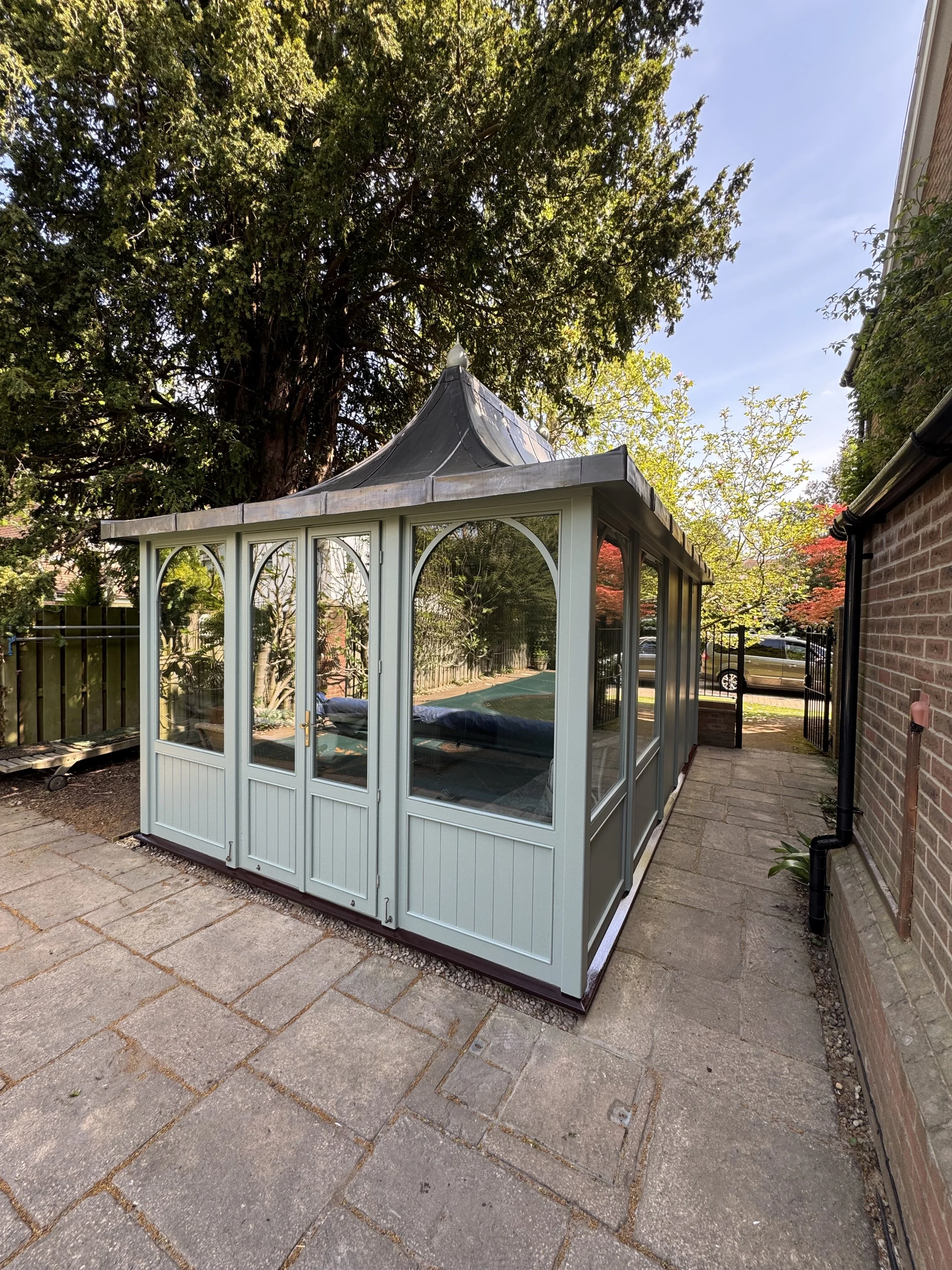 Light green painted garden gazebo with arched window panels and double doors, freshly finished by JN Decorating, set on a paved patio beside a brick house and surrounded by trees.