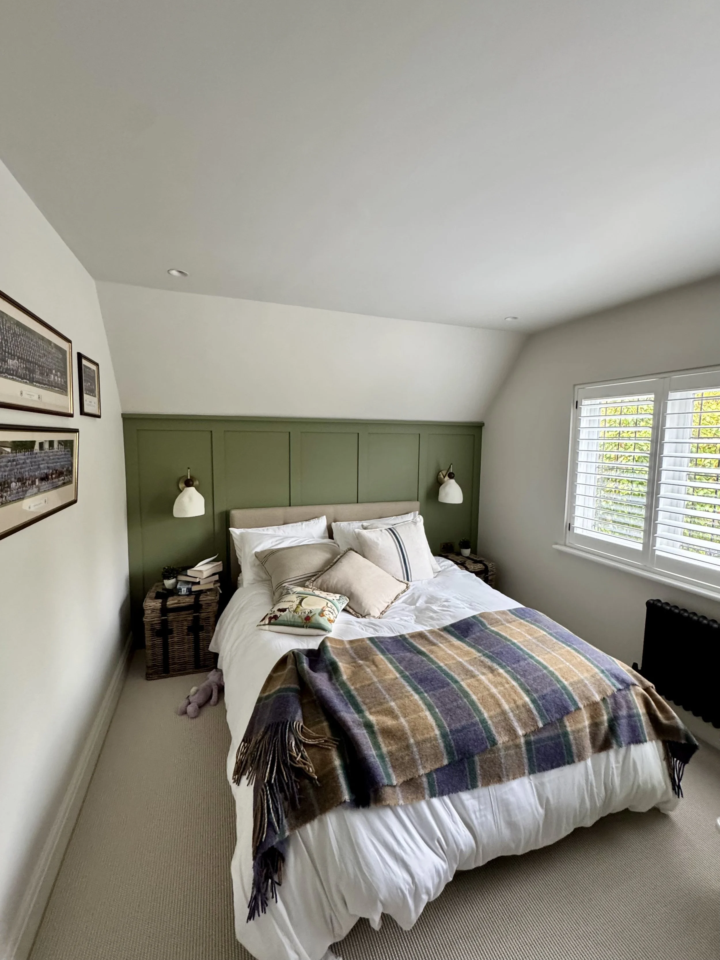 Cozy bedroom with a bed, multiple pillows, a plaid blanket, green accent wall, wall-mounted lamps, side tables, window with plantation shutters, and framed pictures on the wall.