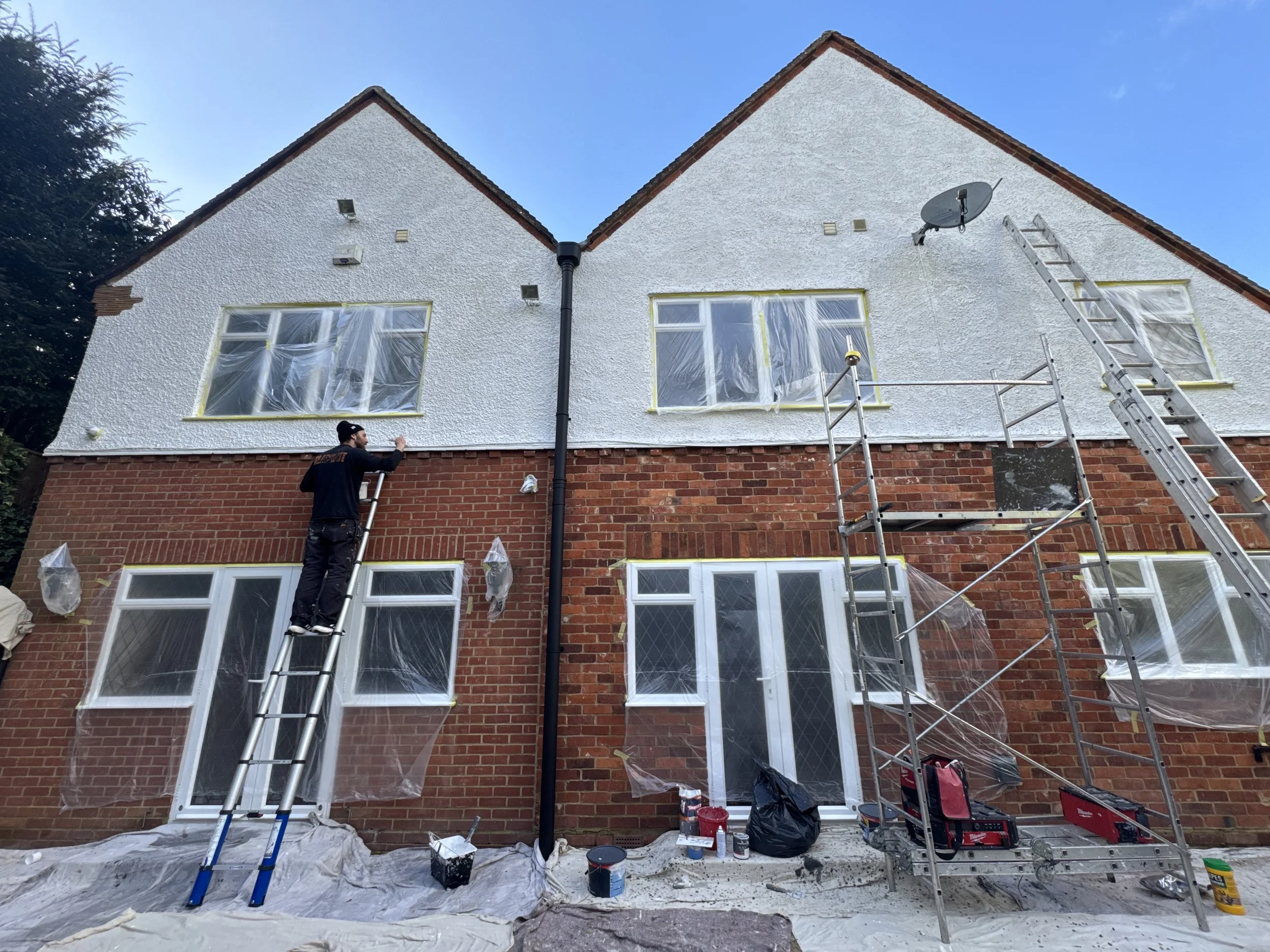 A house is being painted white on the upper walls, with scaffolding and a ladder set up. A man is painting the house from a ladder, and there are paint supplies and plastic coverings around the area.
