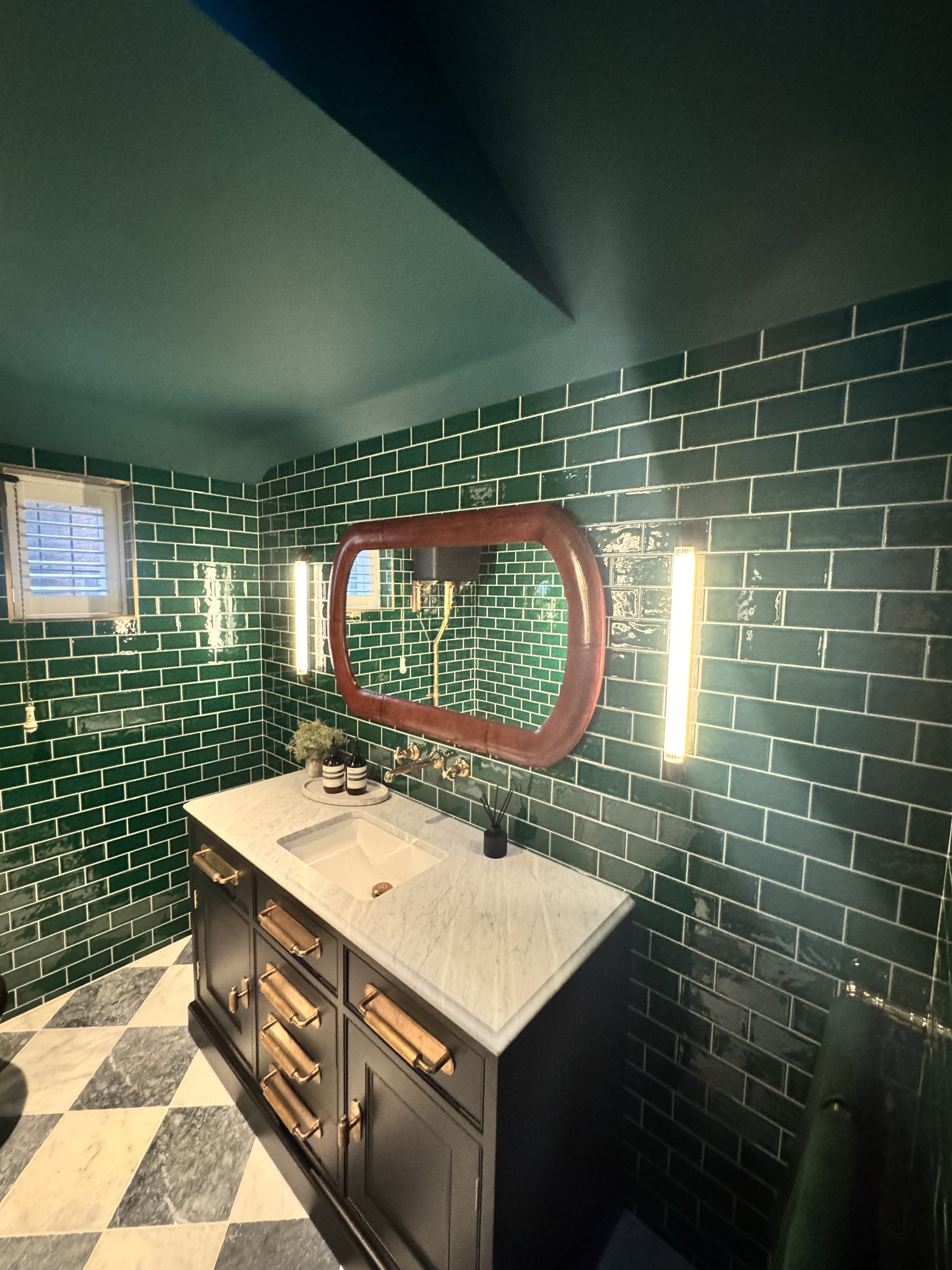 Modern bathroom with dark green subway tile walls, a white marble countertop vanity with a sink, a large mirror with a brown frame, and vertical lighting fixtures on either side of the mirror. There are two small windows with blinds, and the floor has a black, white, and gray checkered pattern.