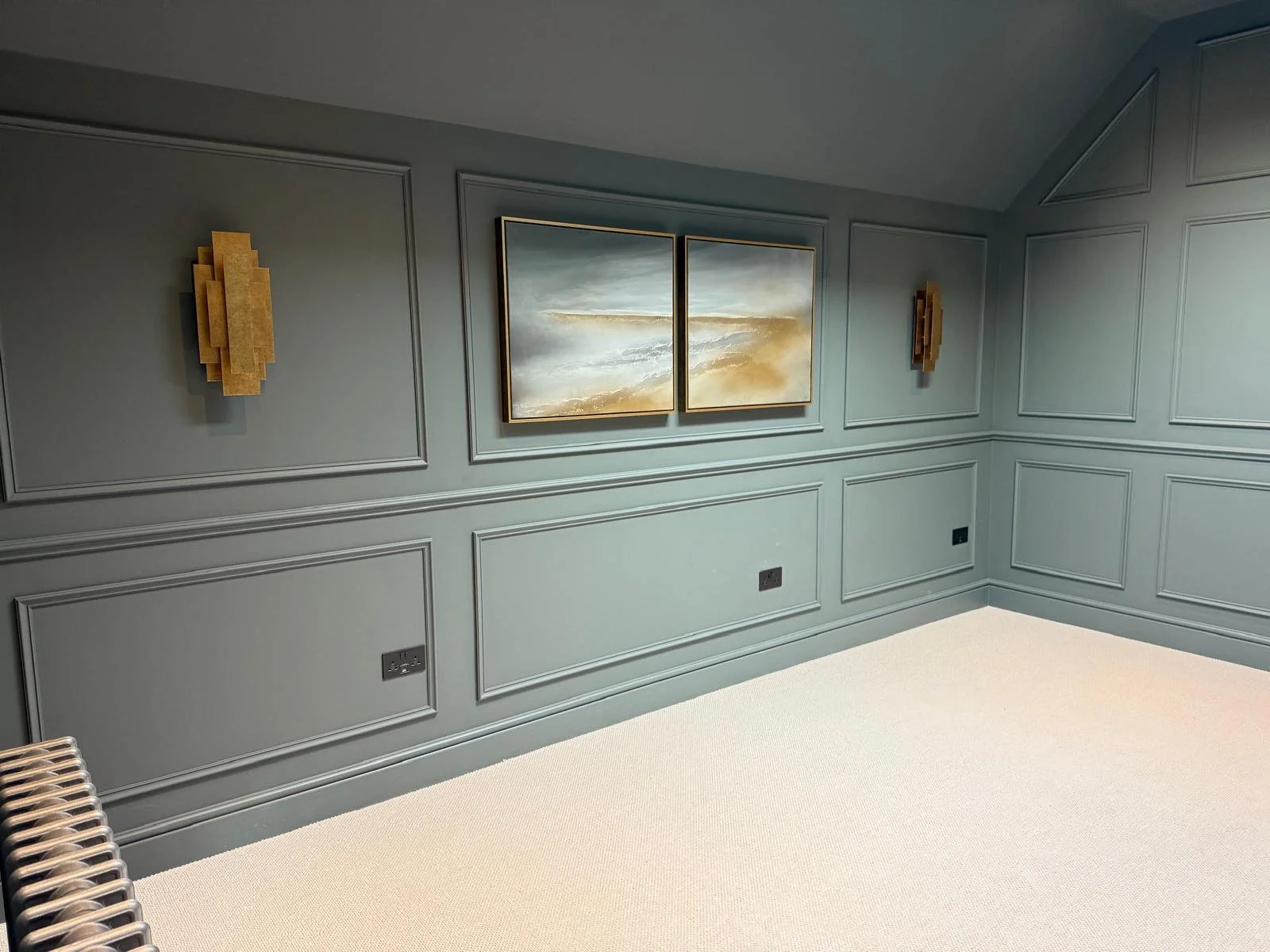Empty room with teal paneled walls, framed abstract art, two gold decorative wall sconces, beige carpet, and electrical outlets.