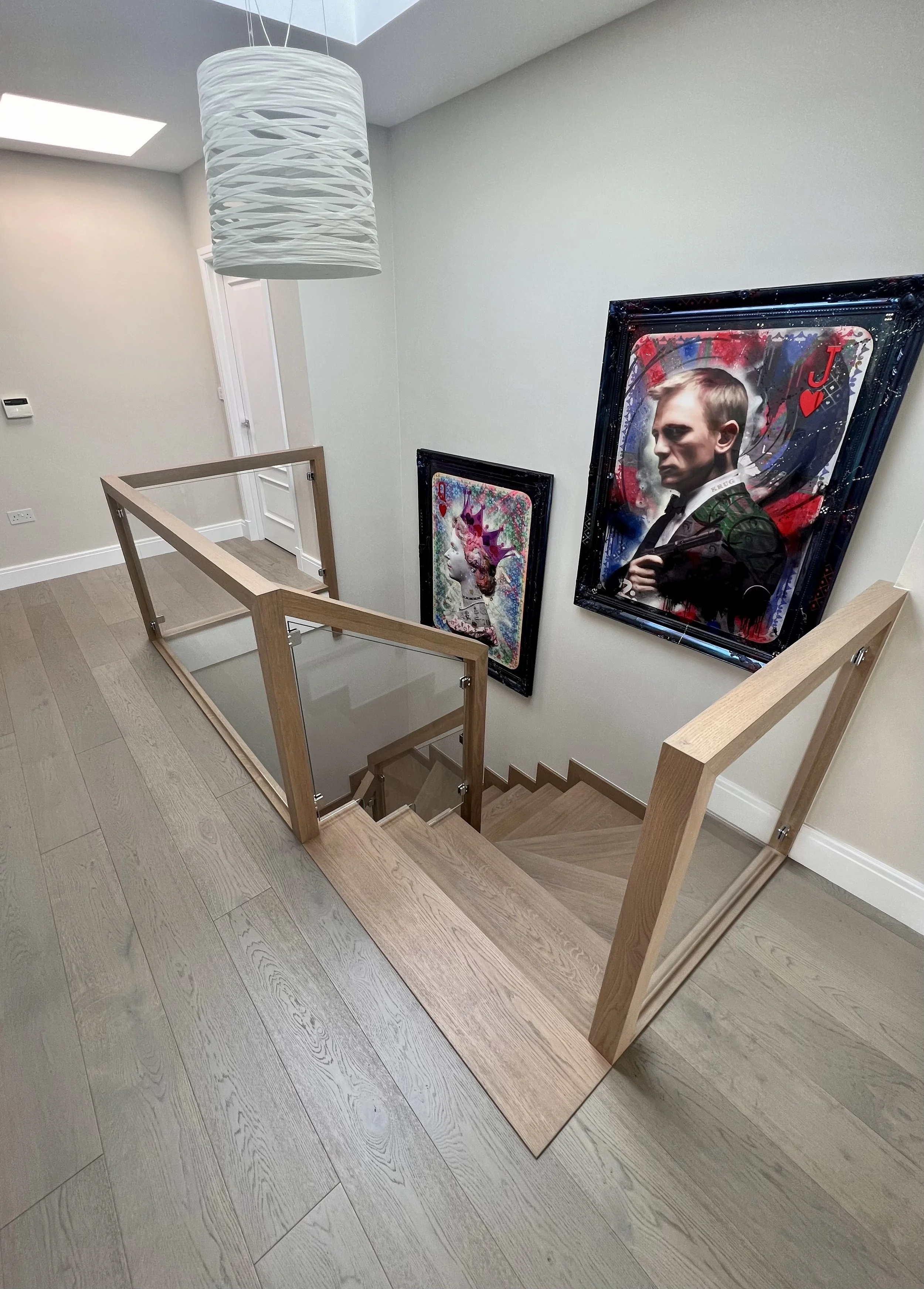 View of a staircase landing with modern artwork of Daniel Craig as James Bond on the wall, a wooden railing with glass panels, hardwood flooring, and a ceiling light fixture.