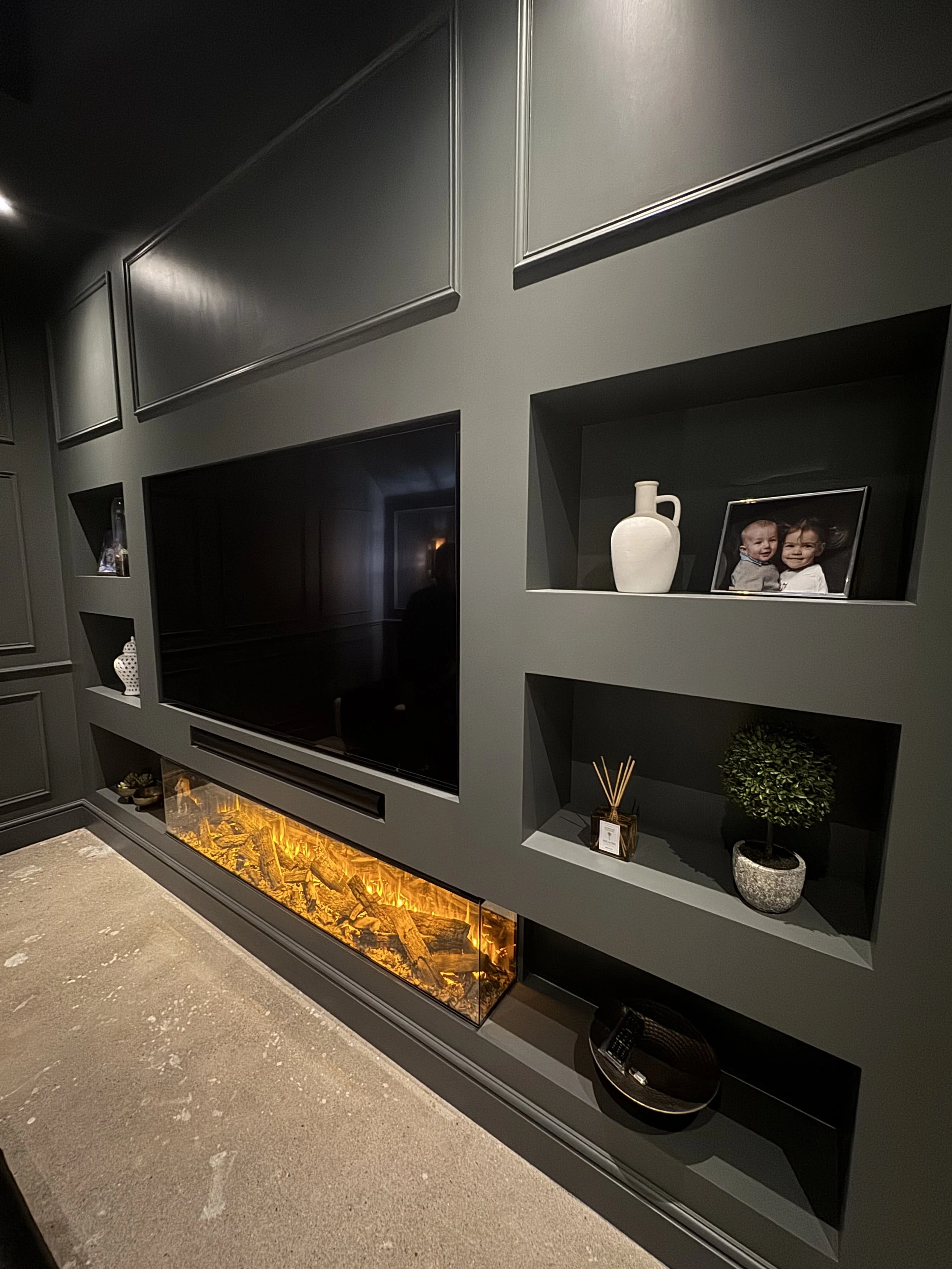 Modern media wall with dark painted panelling, integrated shelving, and a built-in electric fireplace beneath a mounted TV, professionally finished by JN Decorating.