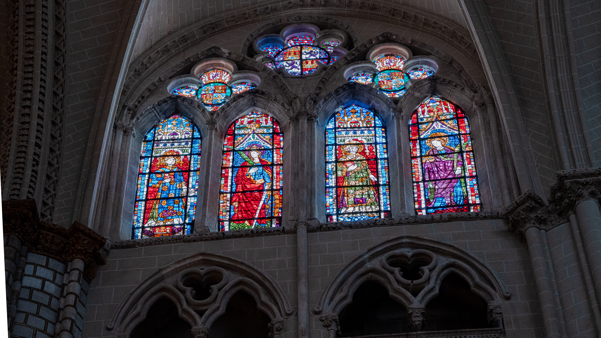 Clerestory of Toledo Cathedral: four stained glass lancets and small rose oculi in deep stone reveals above a blind triforium arcade, the only bright surfaces in the bay