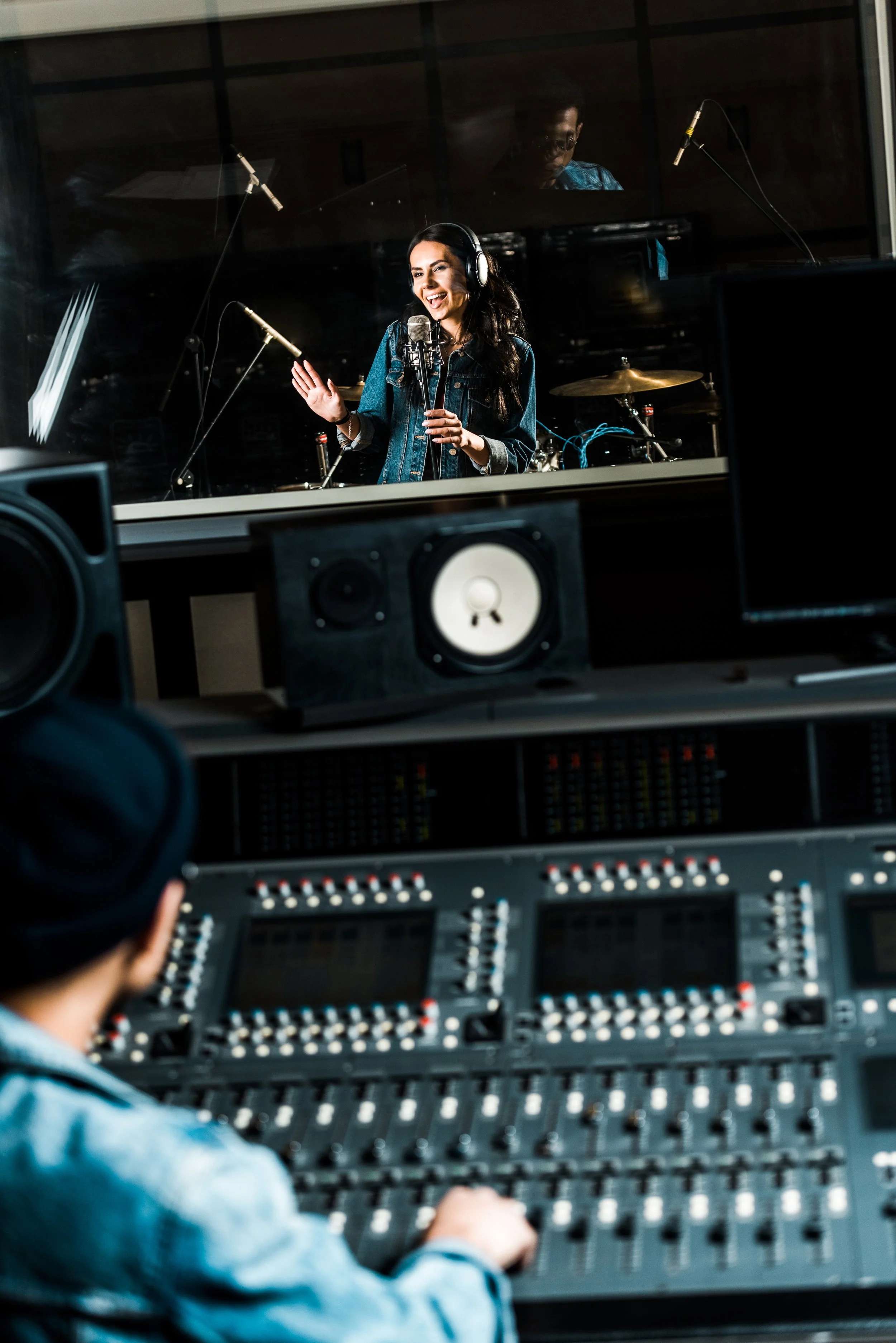 Writing     Singer wearing headphones at a microphone, viewed through a recording studio window, with an audio engineer at the mixing console in the foreground.