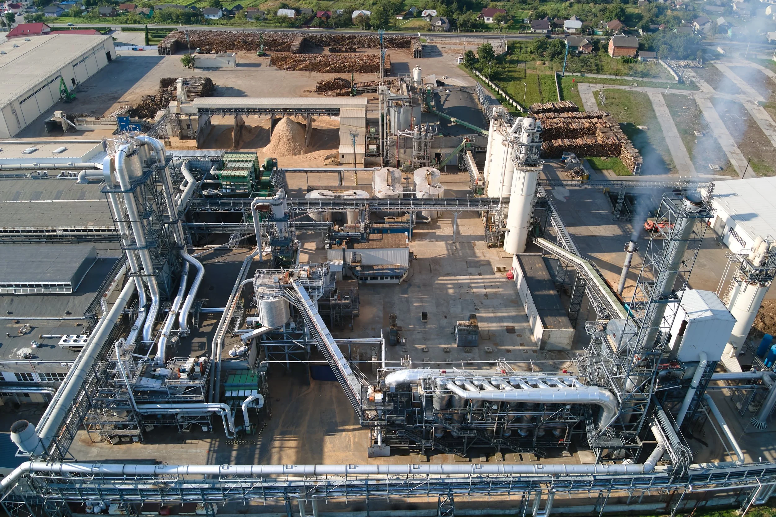 Aerial view of a manufacturing plant with production buildings, silos, extensive pipework, conveyors, and outdoor timber storage.