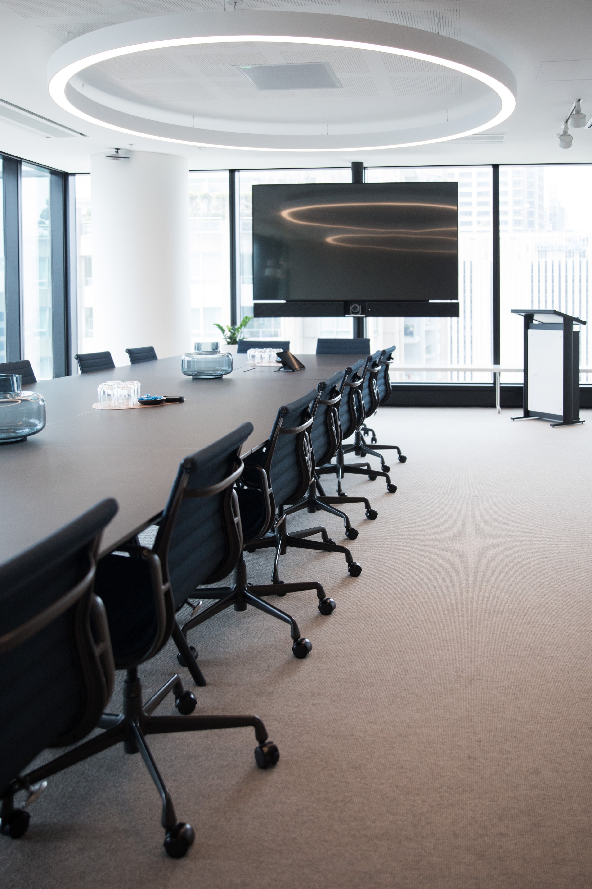 A modern conference room with a large oval table surrounded by black office chairs on wheels, a big flat screen monitor at the end of the room, and floor-to-ceiling windows showing city buildings outside. There is a podium and some decorative items on the table.