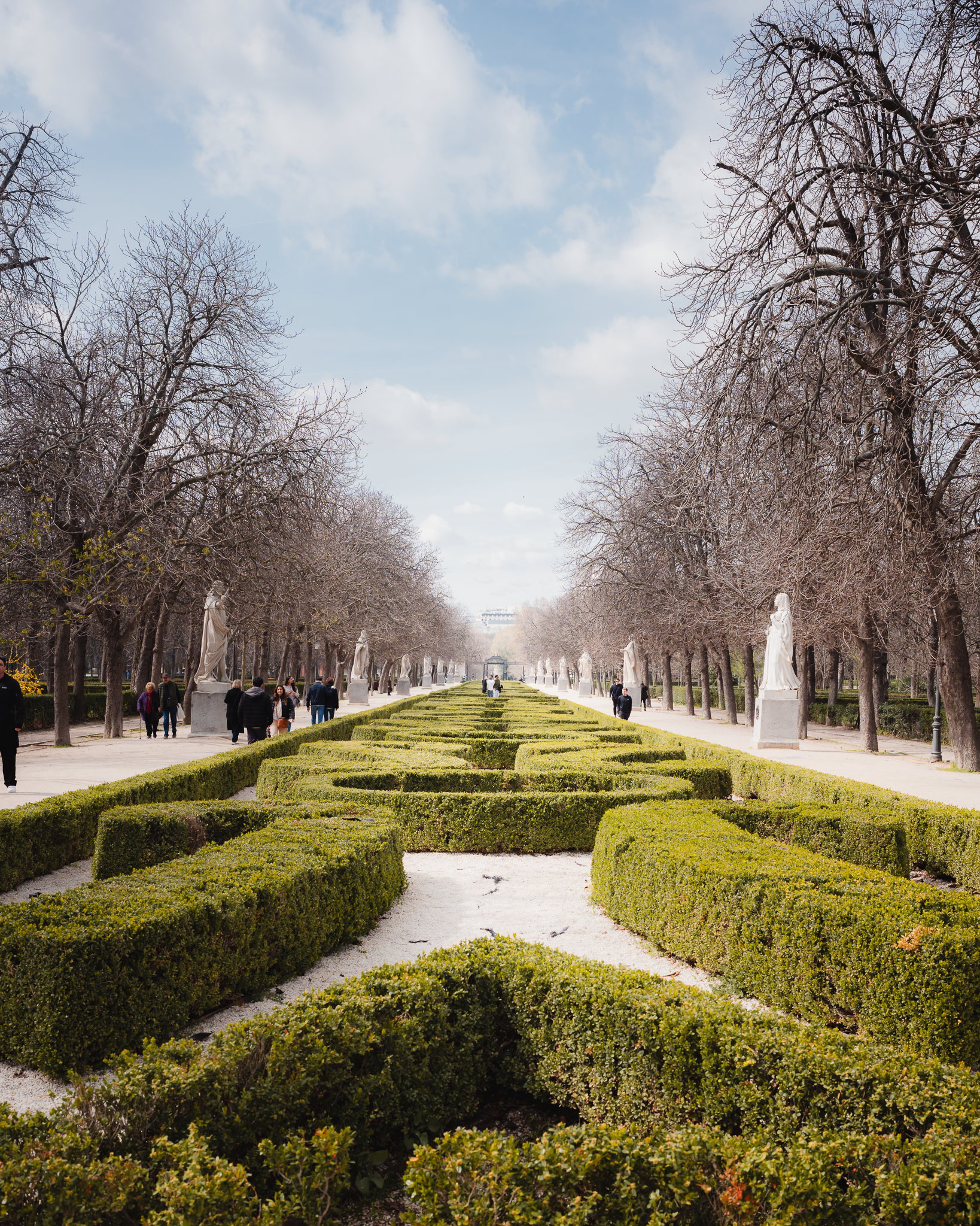Symmetrical clipped boxwood parterre lined with white royal statues along a tree-shaded promenade in El Retiro Park, Madrid