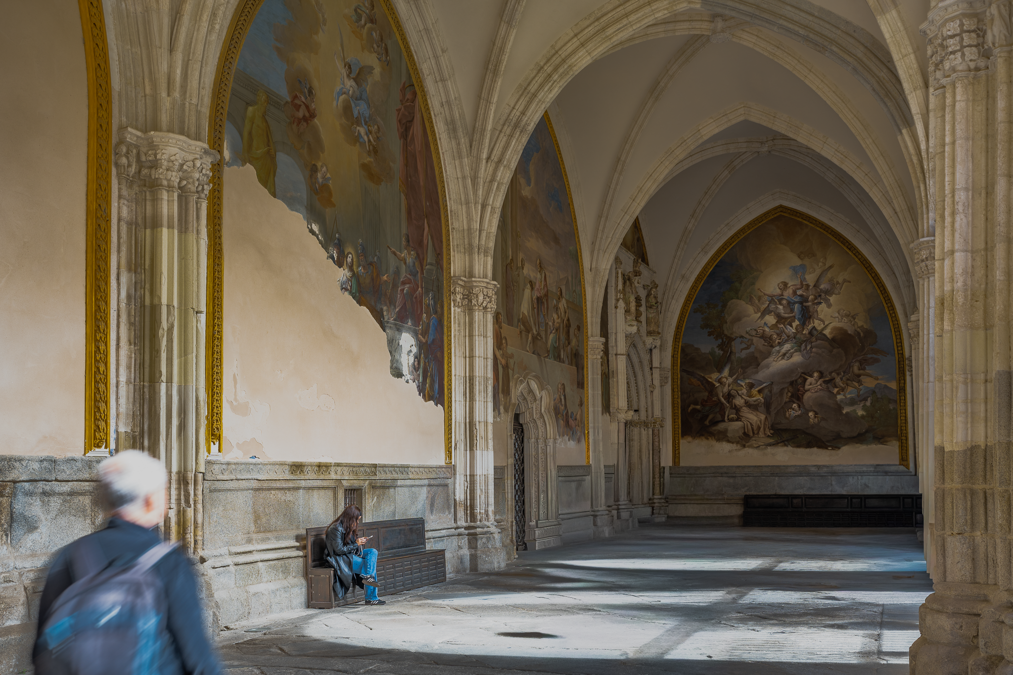 Cloister gallery in Toledo Cathedral lined with painted murals in varying states of loss, with visitors seated on a stone bench and a passer-by in the foreground