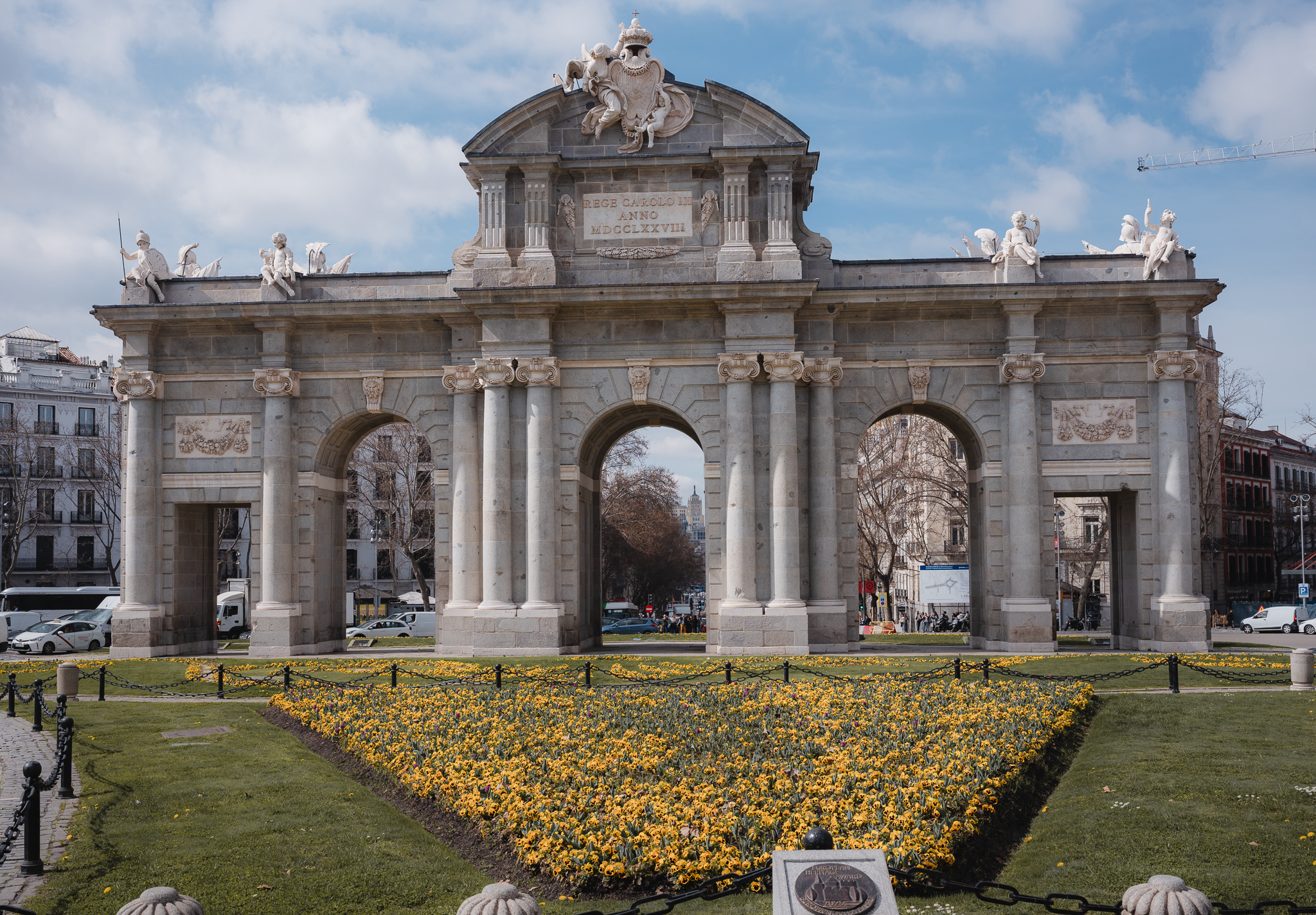 Front view of the Puerta de Alcalá in Madrid behind a triangular bed of yellow spring flowers near El Retiro Park