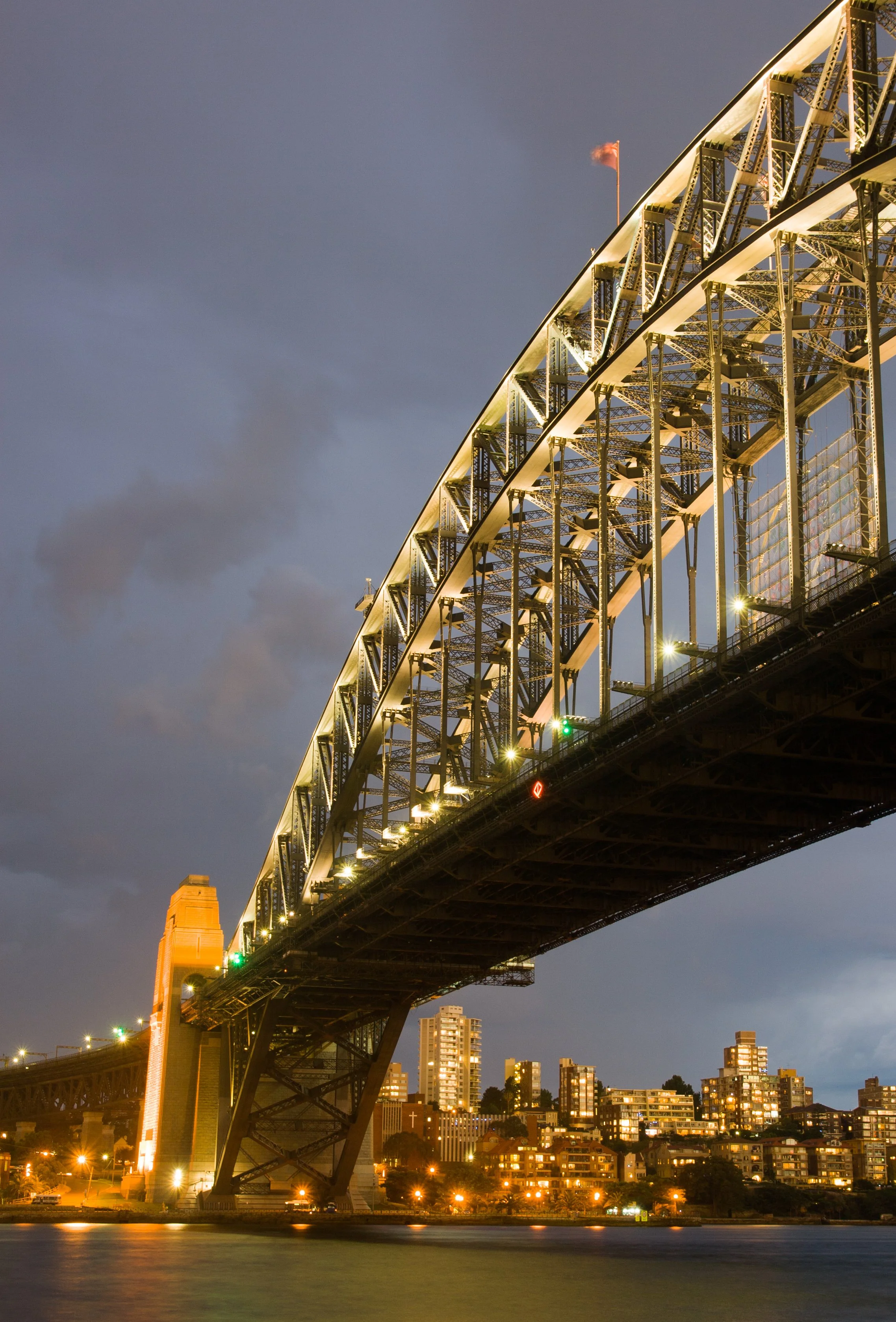 The Sydney Harbour Bridge illuminated at night, reflecting on the water, with city buildings in the background.