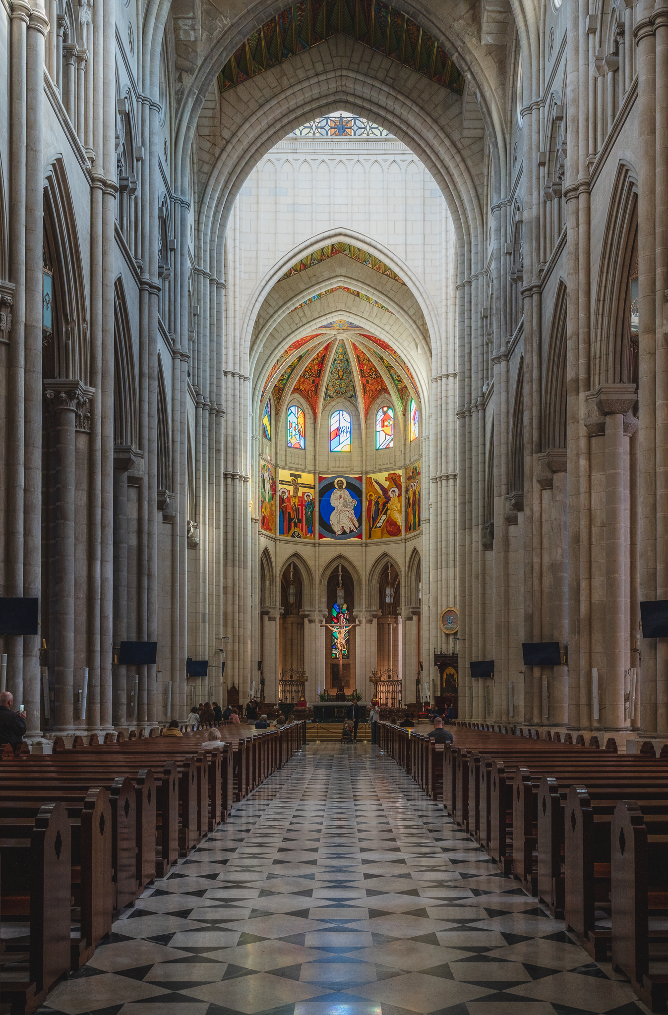 Long central nave of Almudena Cathedral in Madrid, with pointed stone arches, patterned floor, pews and the brightly coloured sanctuary beyond.