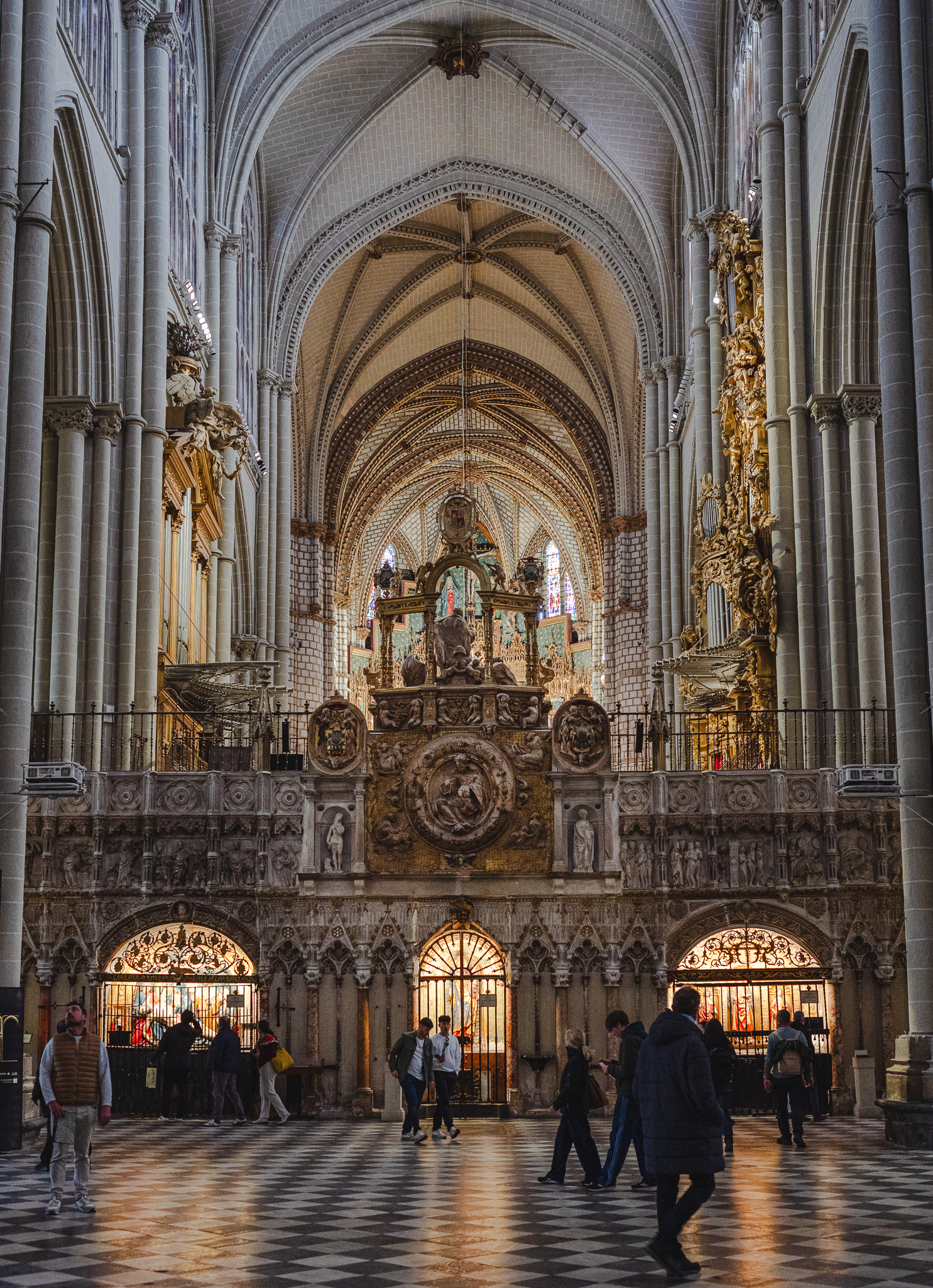 Visitors crossing the chequered marble floor of Toledo Cathedral's nave in front of the carved rear screen of the choir, with daylight reflecting off the polished stone