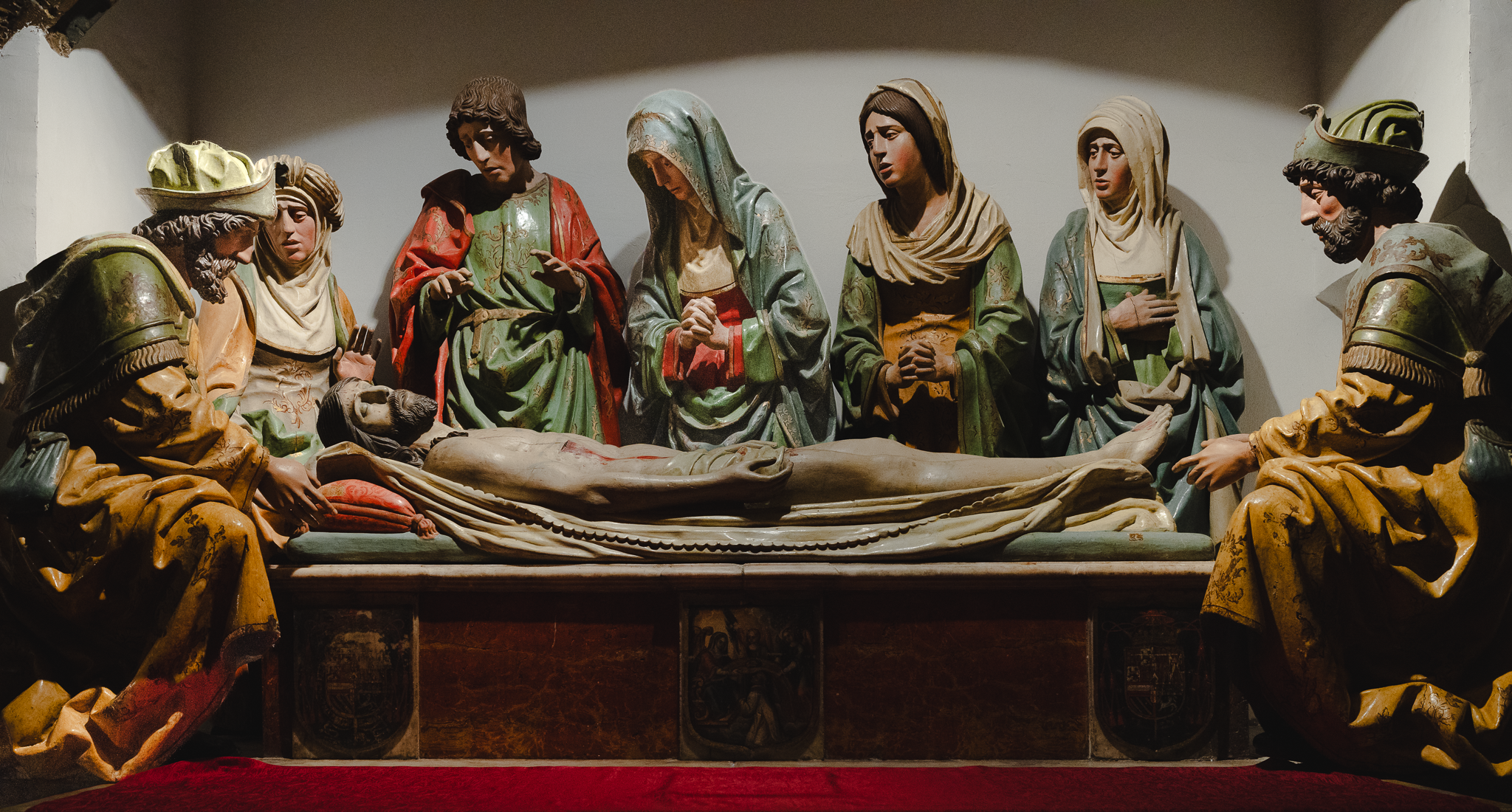Polychrome sculpted entombment group in Toledo Cathedral, mourning figures in green, red and gold robes around a recumbent figure, lit by directional museum lighting