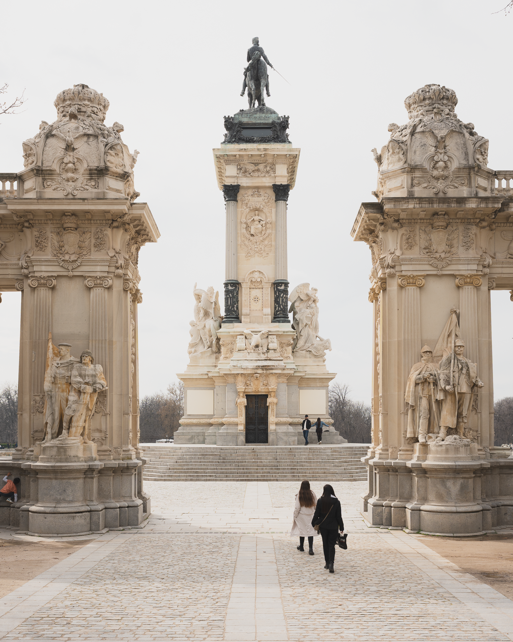 Two visitors walking toward the Alfonso XII monument in El Retiro Park, Madrid, framed by sculpted colonnade pavilions
