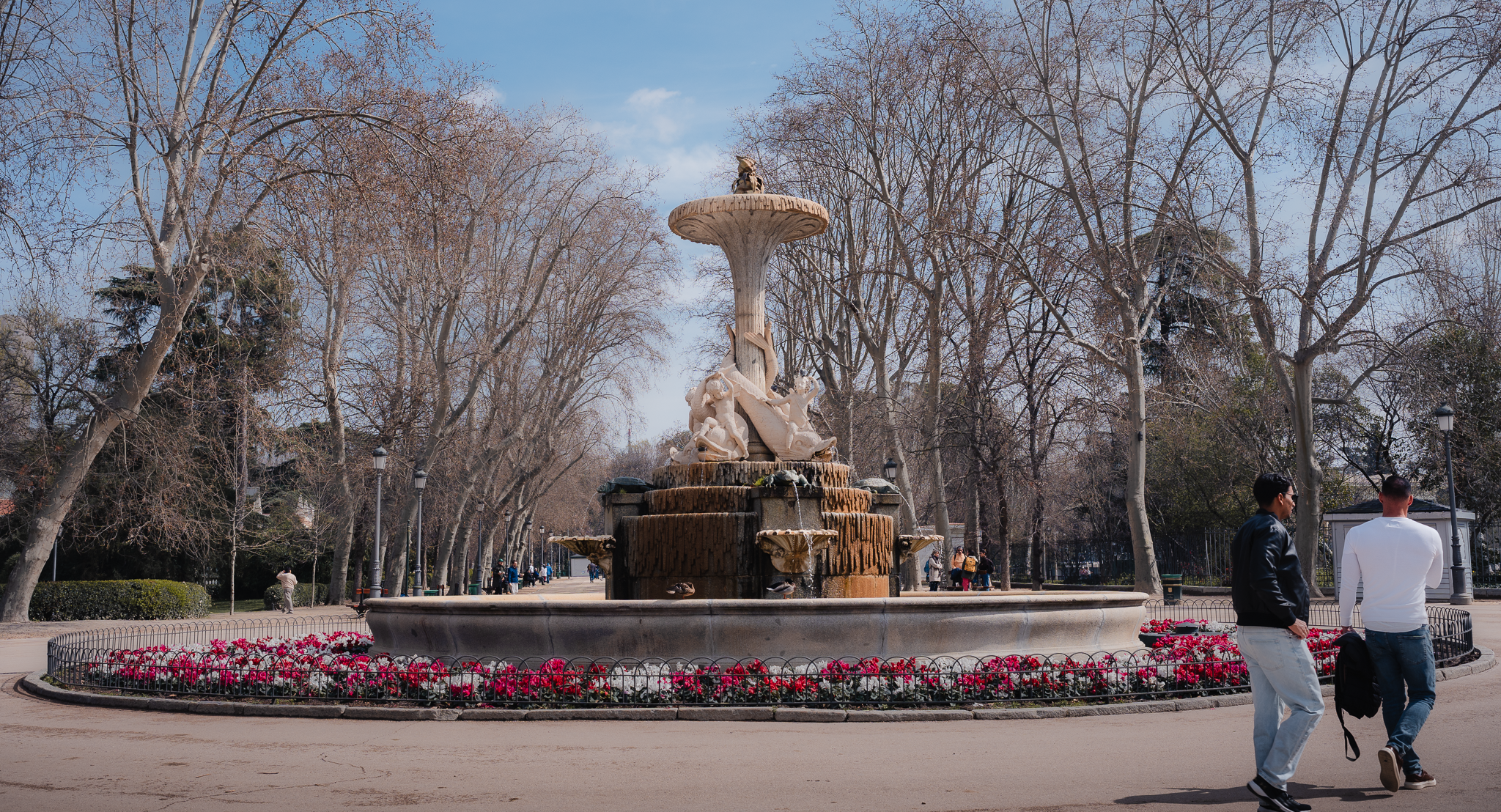 Fuente de la Alcachofa (Artichoke Fountain) in El Retiro Park, Madrid, ringed by pink flowers as two men walk past