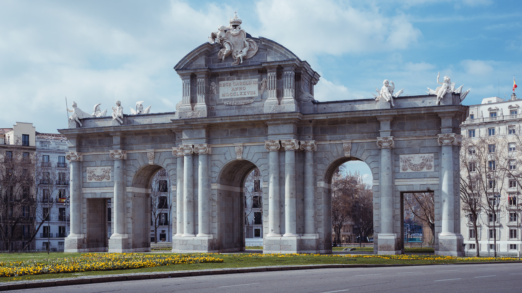 Puerta de Alcalá neoclassical stone gateway in Madrid, framed by a bed of yellow spring flowers under a blue sky