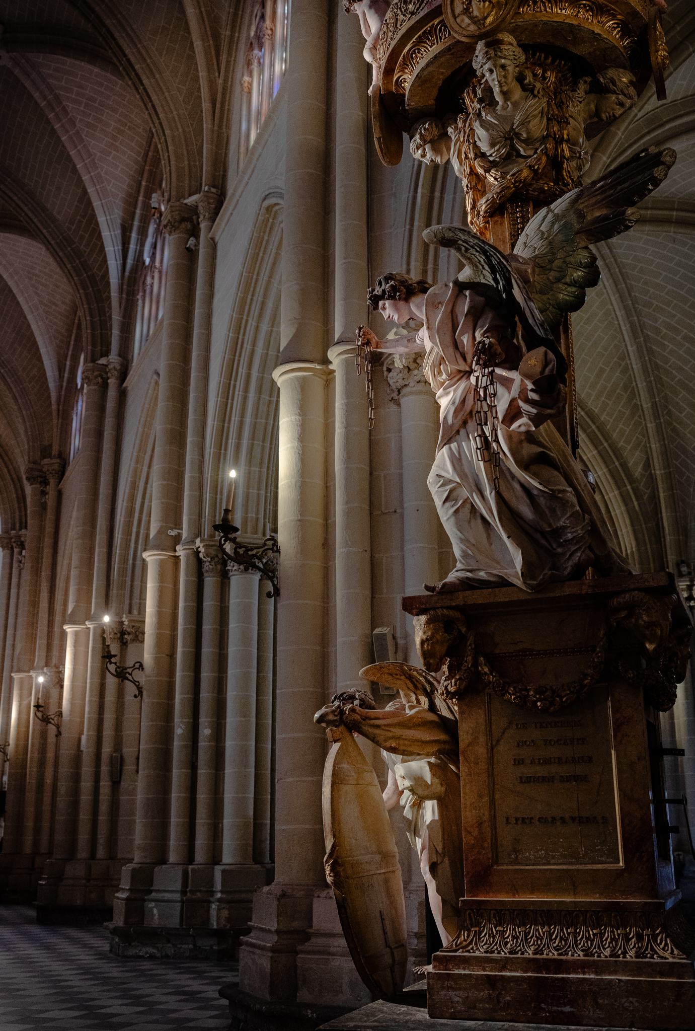 Winged angel carved on Toledo Cathedral's paschal candlestick, seen against the receding side aisle with its stone piers and lit wall sconces