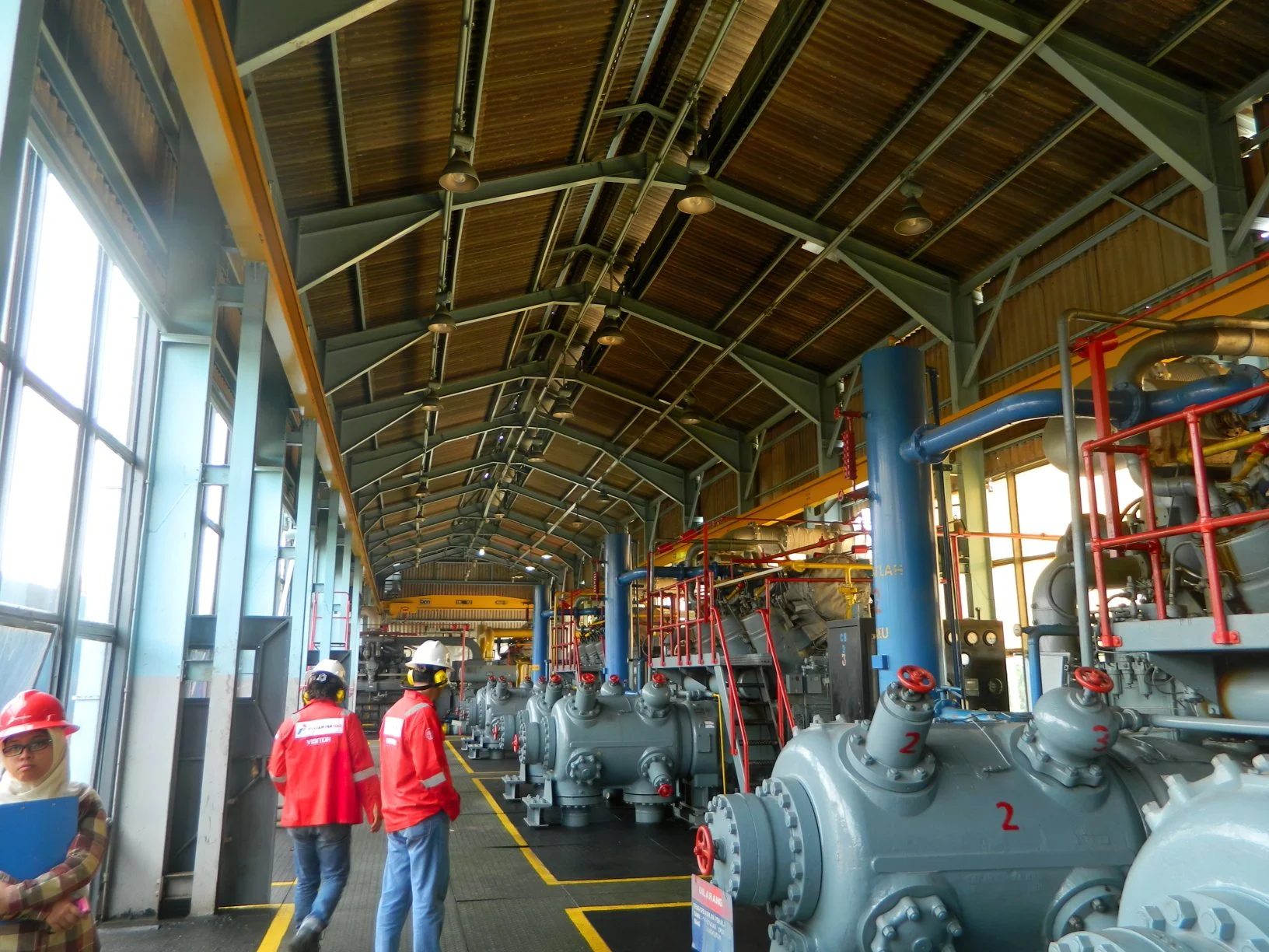 Natural Gas facility interior with large machinery, pipework, and personnel wearing hard hats beside a marked walkway.