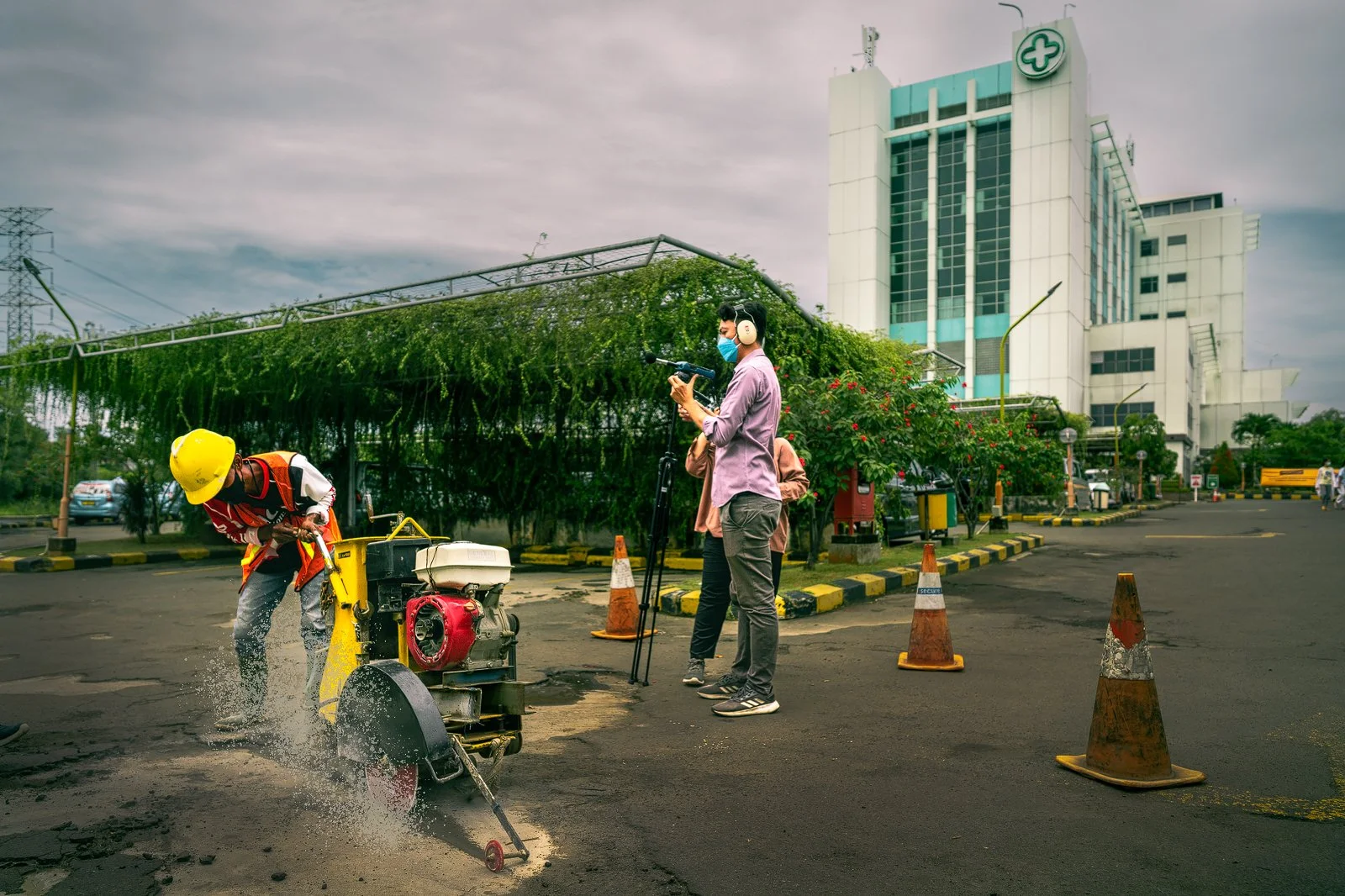 Construction noise measurement beside a worker operating a pavement saw, with a hospital building in the background.