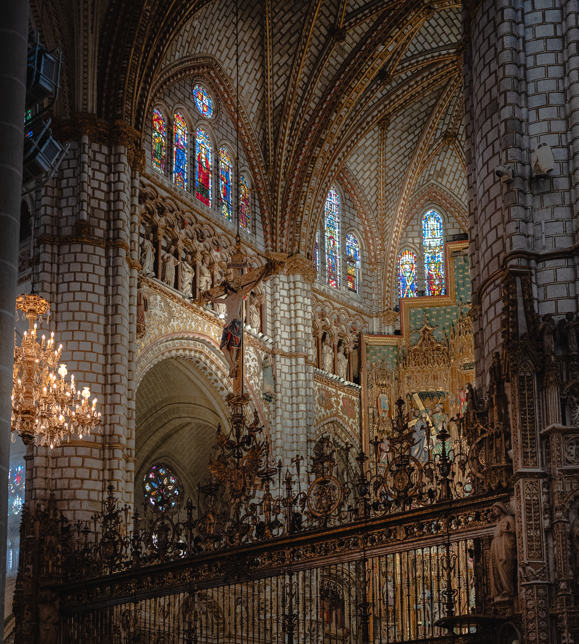 Upper level of Toledo Cathedral seen from the choir: stained glass lancets, a sculpted triforium gallery, patterned ribbed vaults, a suspended crucifix and a lit chandelier