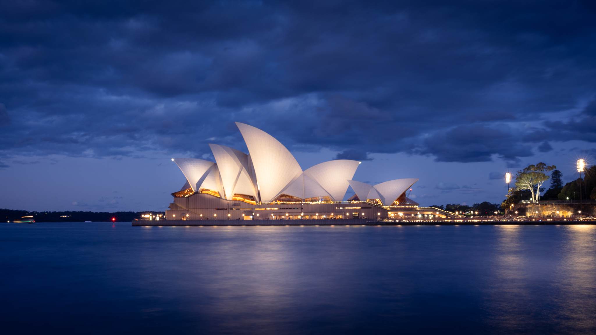 Sydney Opera House illuminated at night with a cloudy sky and water in the foreground.