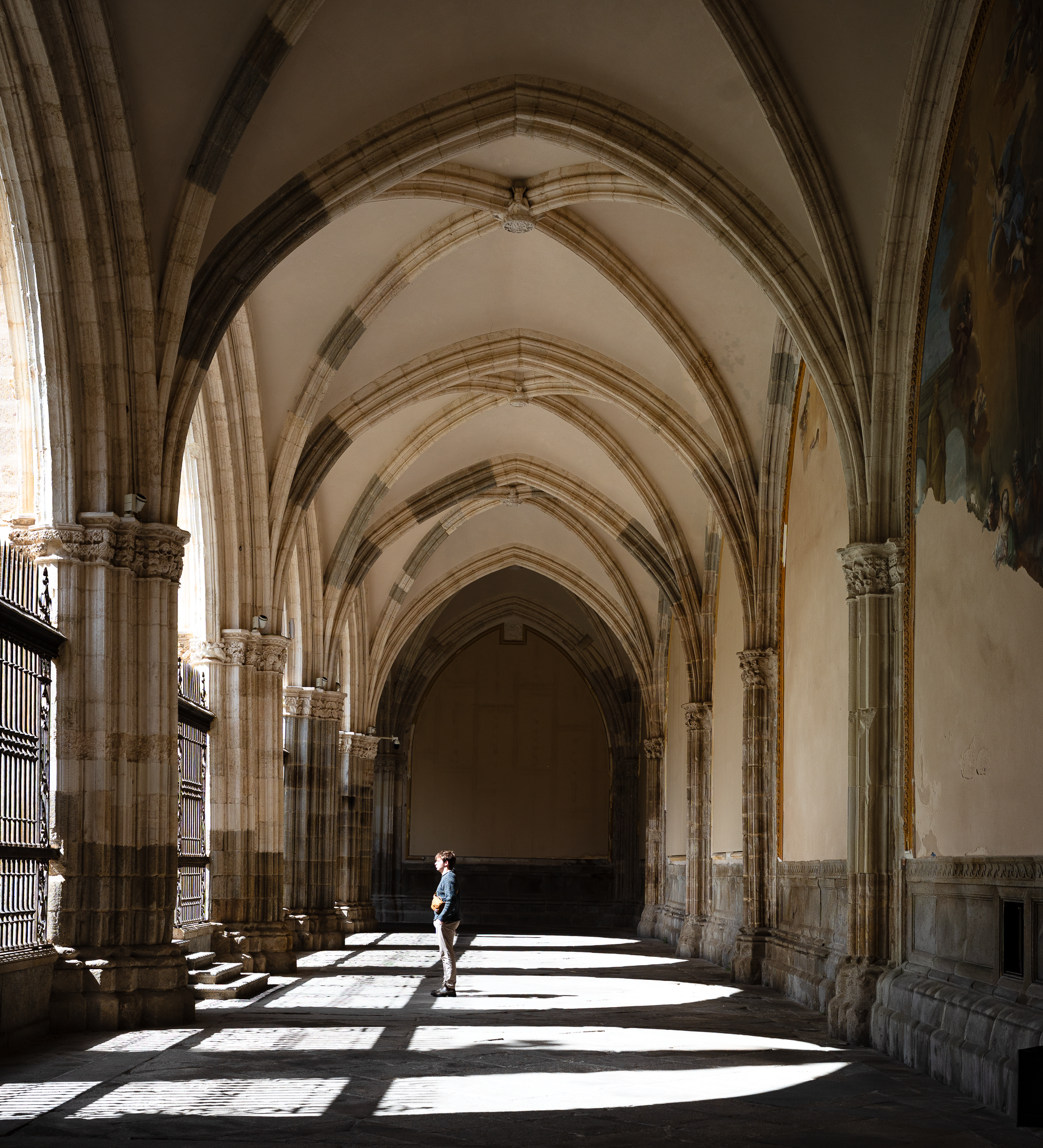 Vaulted cloister walk in Toledo Cathedral with sunlight cast in bright window-shaped patches across the stone floor, a single visitor standing in the light
