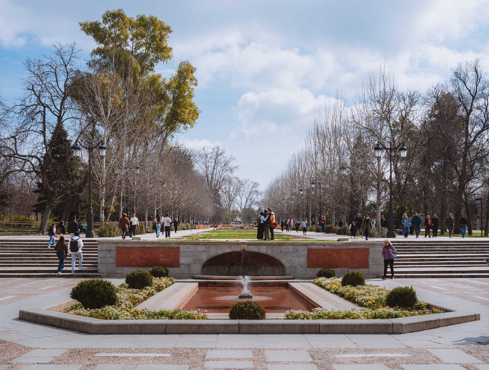 Stone fountain and terraced steps at the Puerta de España entrance to El Retiro Park, Madrid, with visitors walking on a spring afternoon