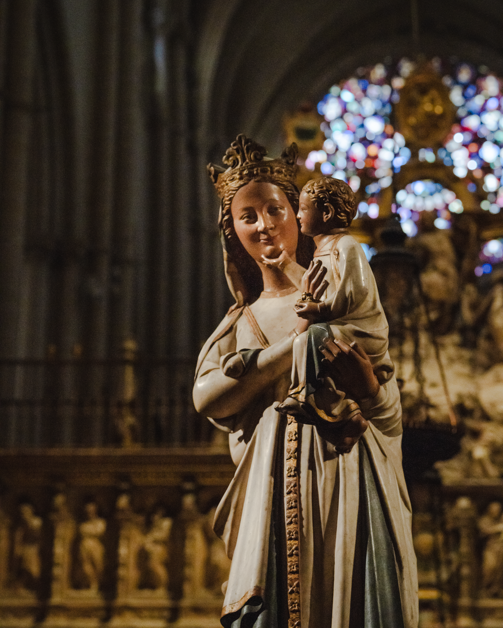 Polychrome sculpture of the Virgin and Child in Toledo Cathedral, sharply focused against a blurred stained glass rose window and carved screen behind