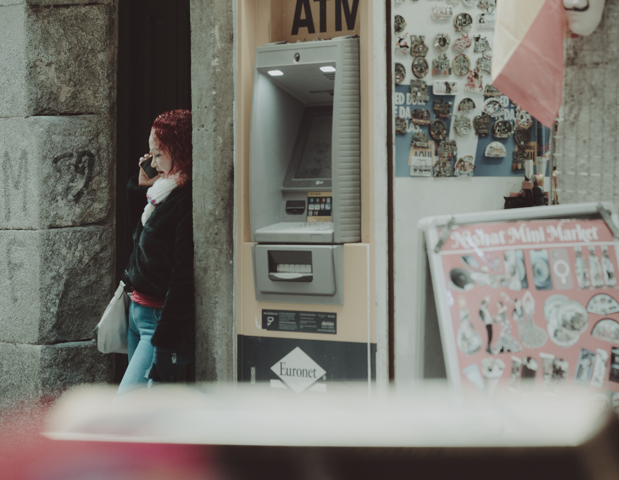 Woman with red hair talking on her phone beside a Euronet ATM and a souvenir shop display on a Madrid street