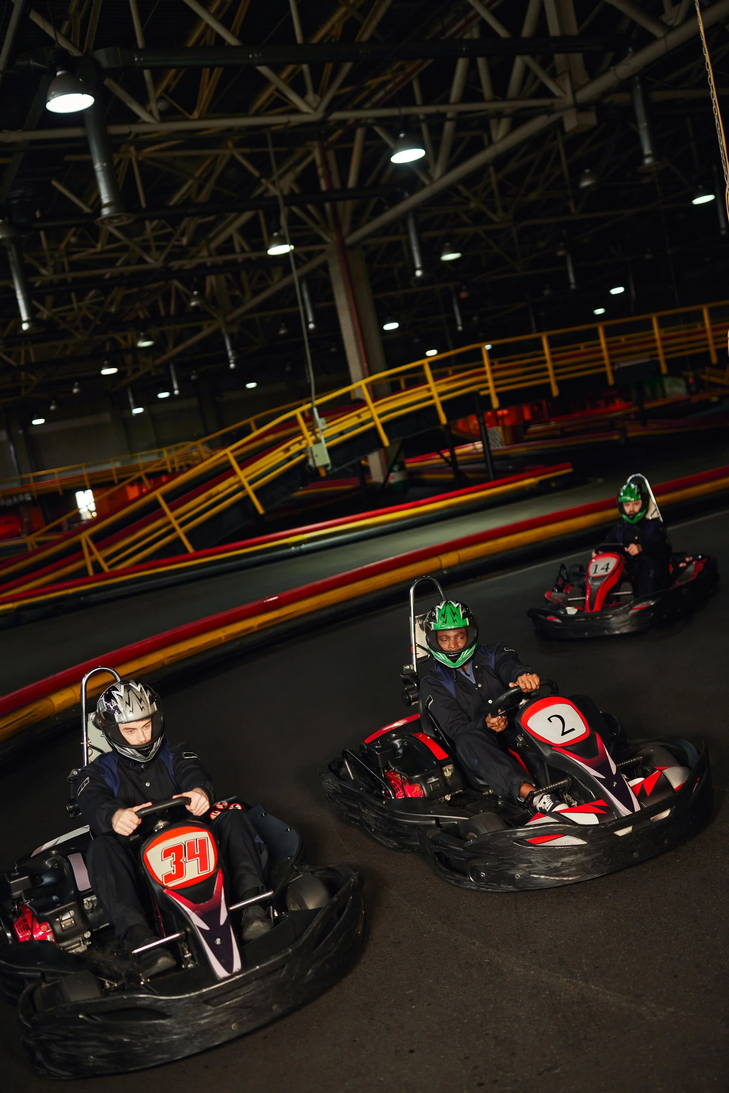 Indoor go-kart race track with three people wearing helmets and racing suits, driving black go-karts with red and white accents, numbered 34, 2, and 14. The track has colorful barriers and overhead lighting.