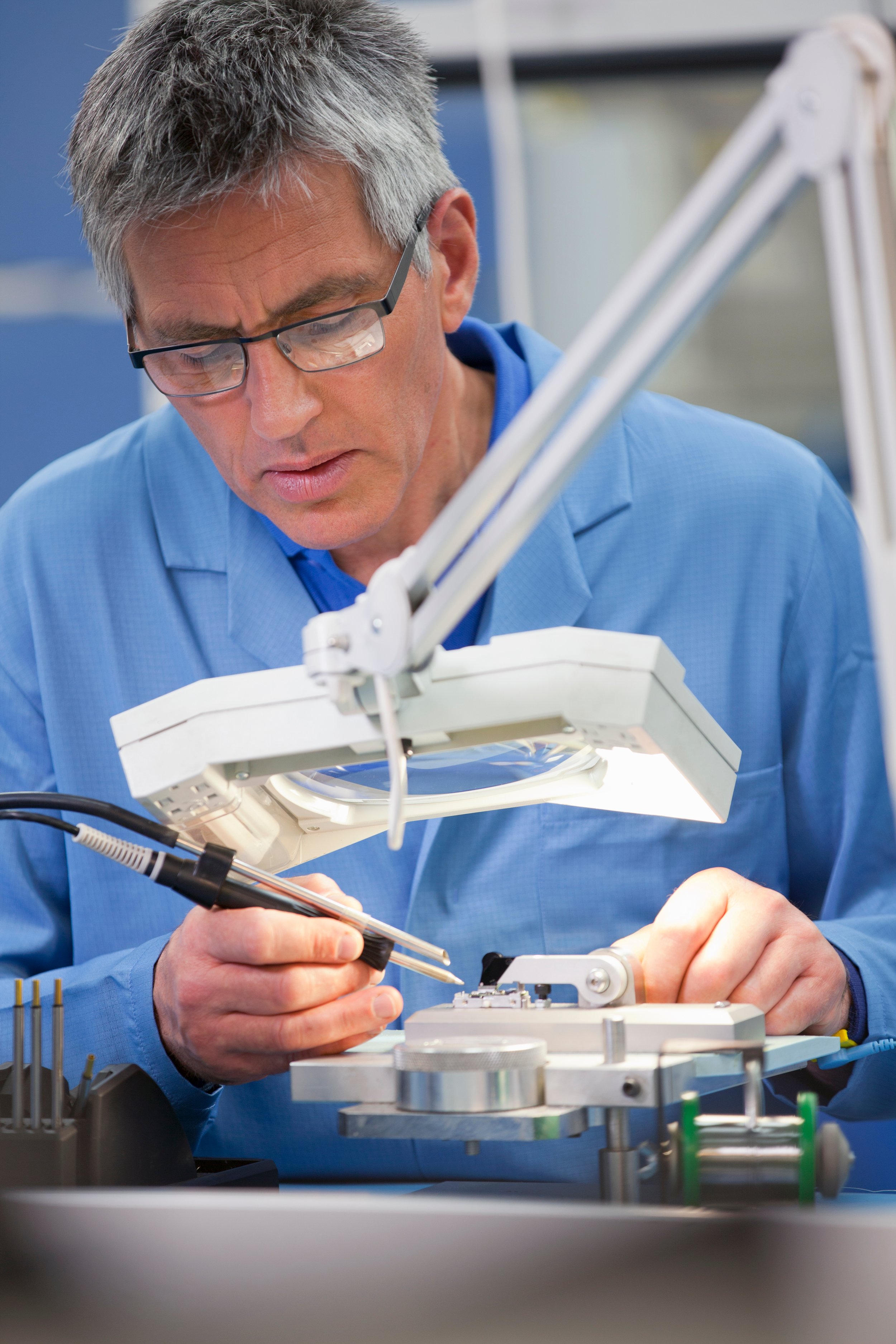 A scientist wearing glasses and a blue lab coat working with scientific equipment in a laboratory, examining or assembling a device under a bright light.