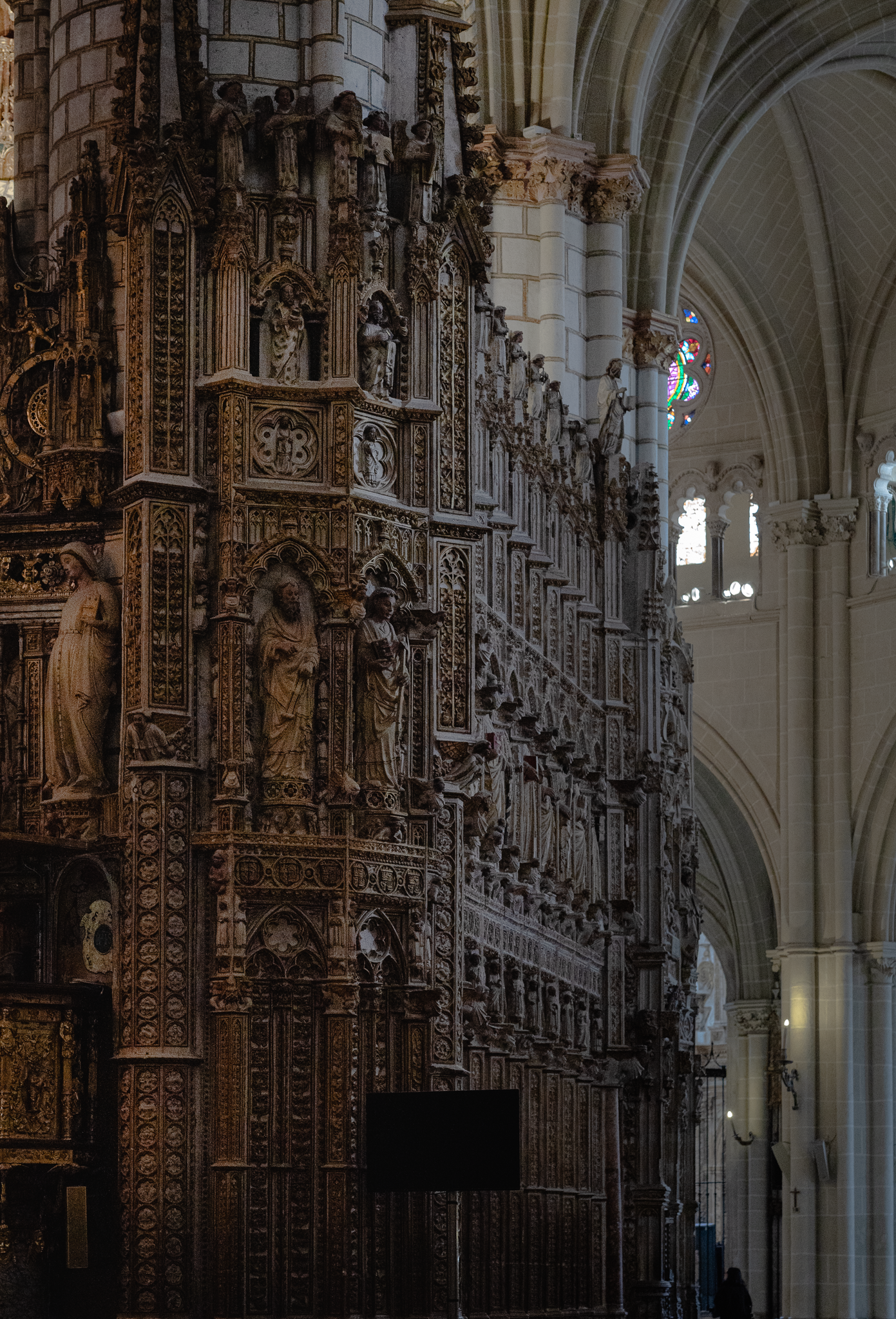 Deeply carved dark stone choir enclosure in Toledo Cathedral set against the pale limestone nave piers and ribbed vault beyond, with a stained glass window glowing in the shadowed aisle
