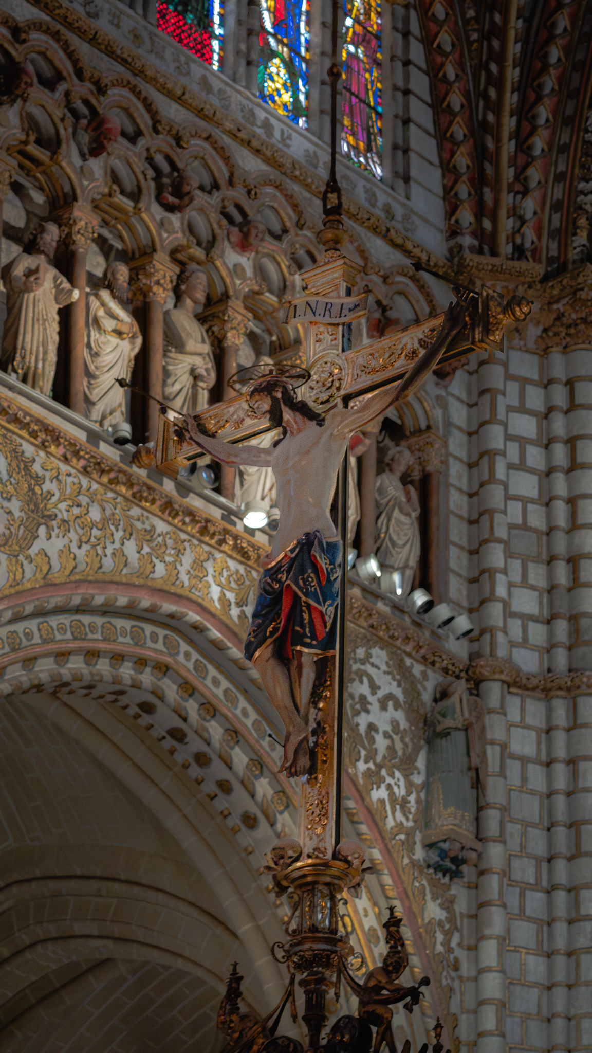 Polychrome crucifix suspended above the nave of Toledo Cathedral, with a statue-filled triforium arcade, stained glass clerestory and modern track-mounted spotlights behind