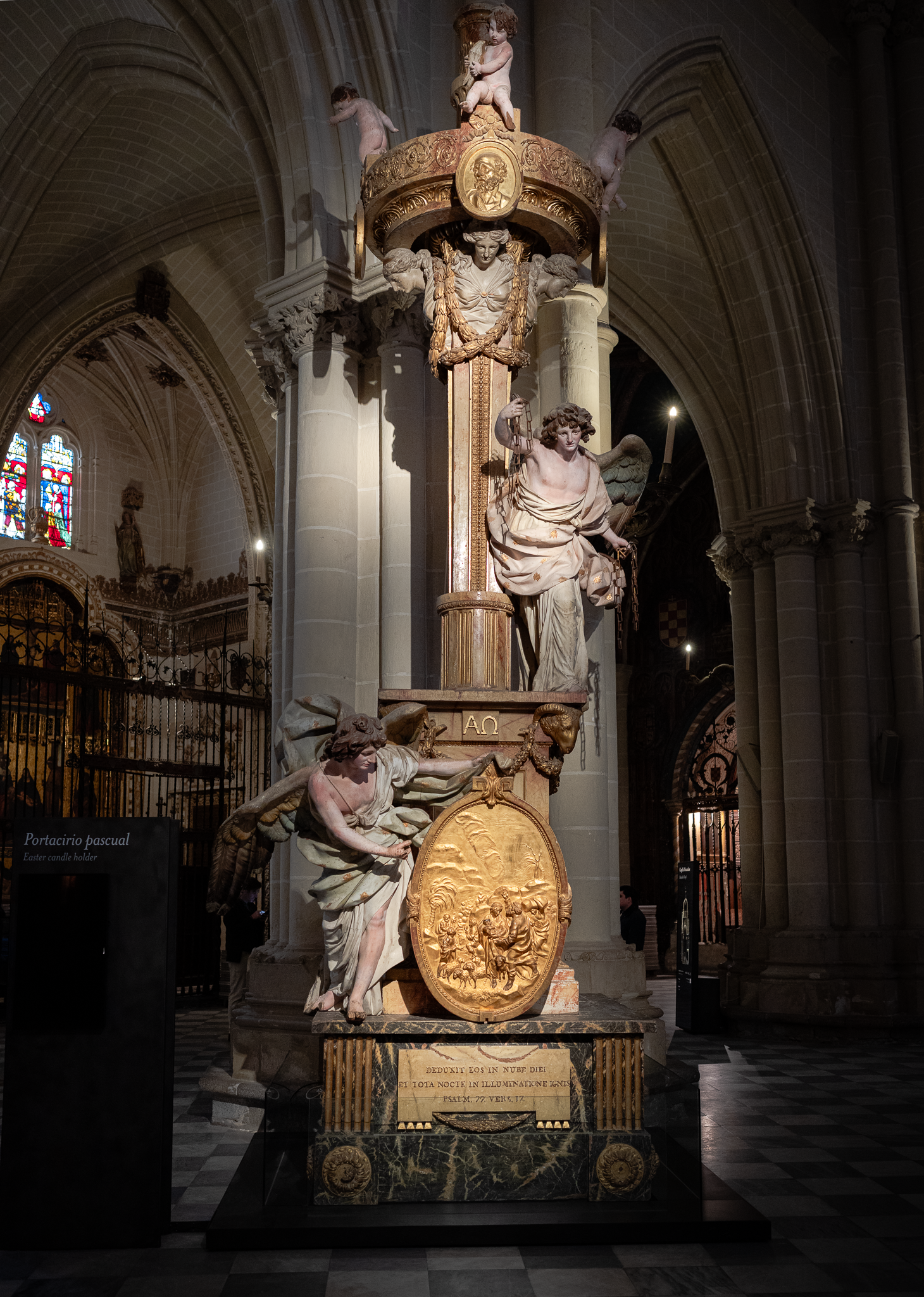 Marble and gilt paschal candlestick in Toledo Cathedral, carved with kneeling angels and a gilded relief medallion, spotlit against the dim aisle behind