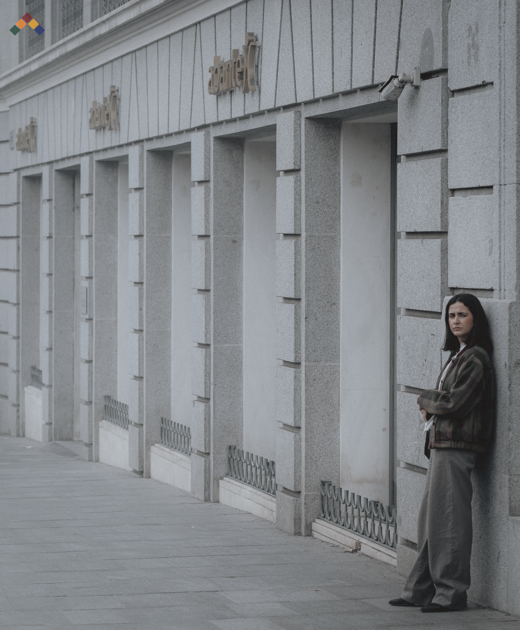 Woman in a brown bomber jacket leaning against the grey stone façade of an Abanca bank building in Madrid