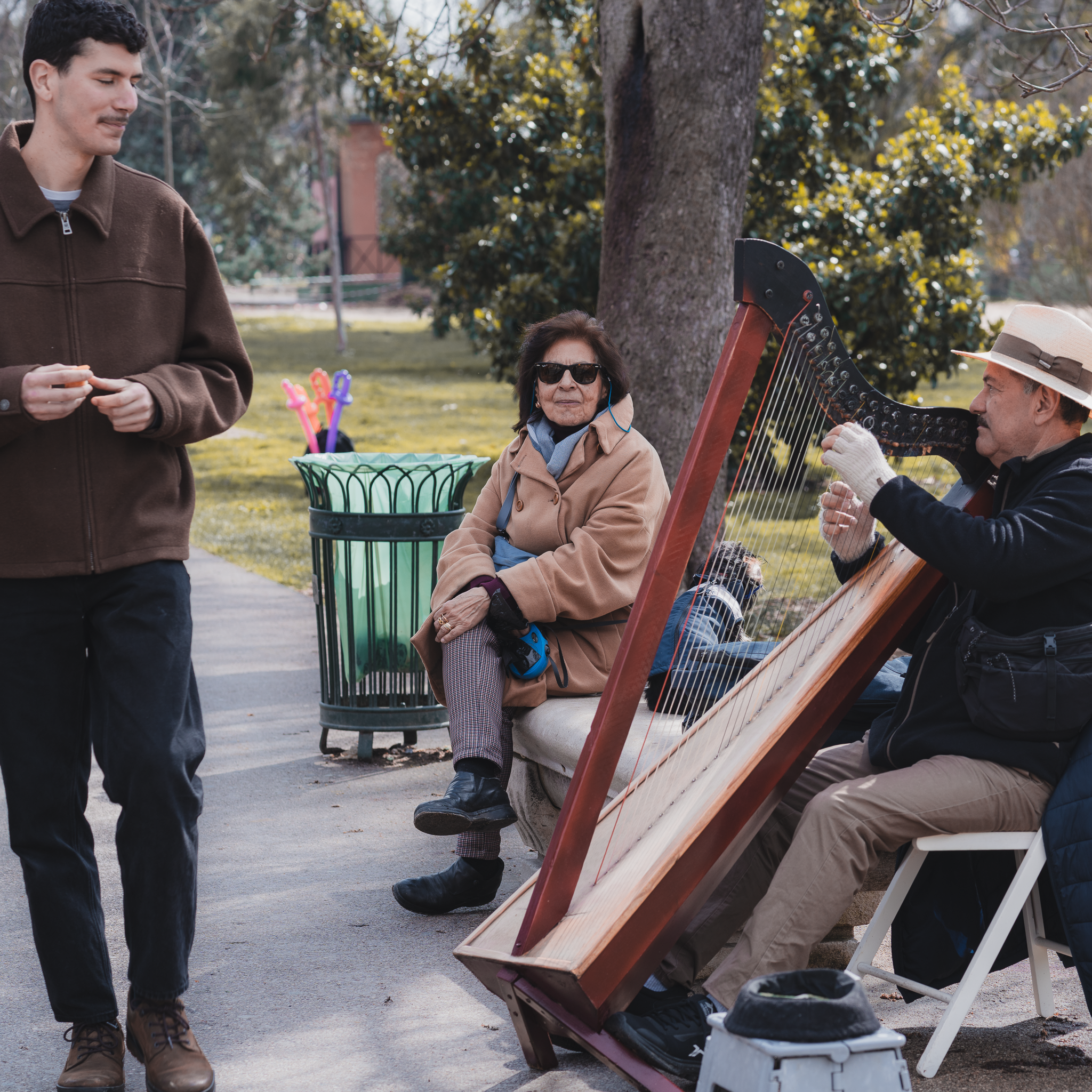 Elderly harpist busking in El Retiro Park, Madrid, as a seated woman and a passing man listen