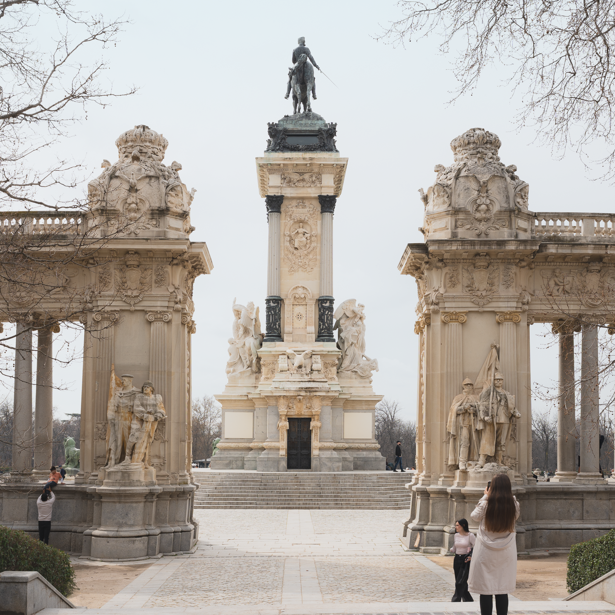 Monument to Alfonso XII in El Retiro Park, Madrid, seen between two ornate stone pavilions on an overcast day