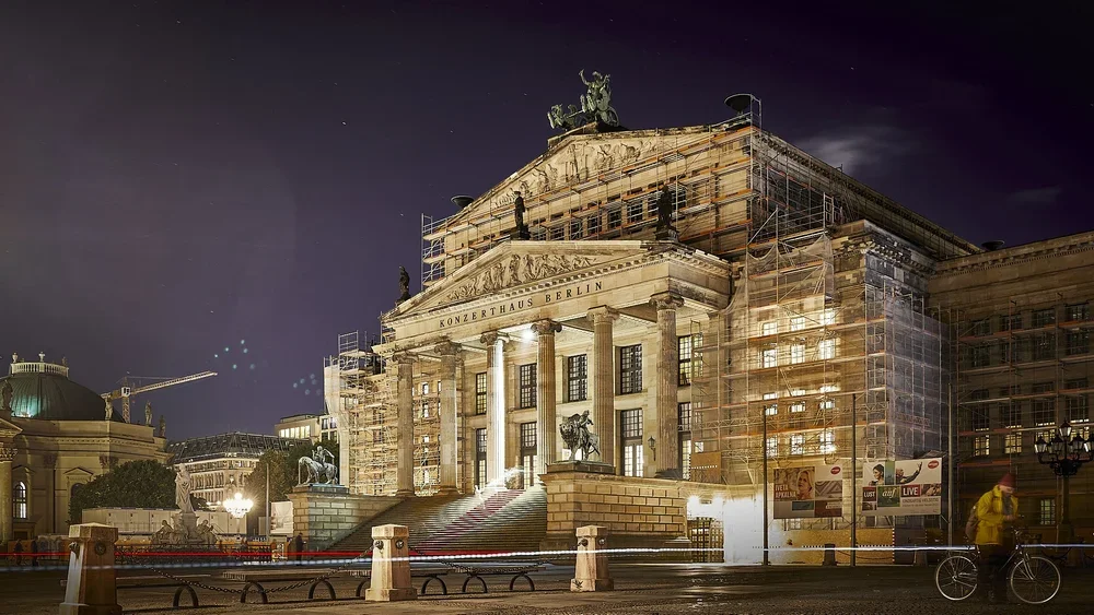 Konzerthaus Berlin seen at an angle from Gendarmenmarkt at night, with broad entrance steps, tall columns, and scaffolding across the façade