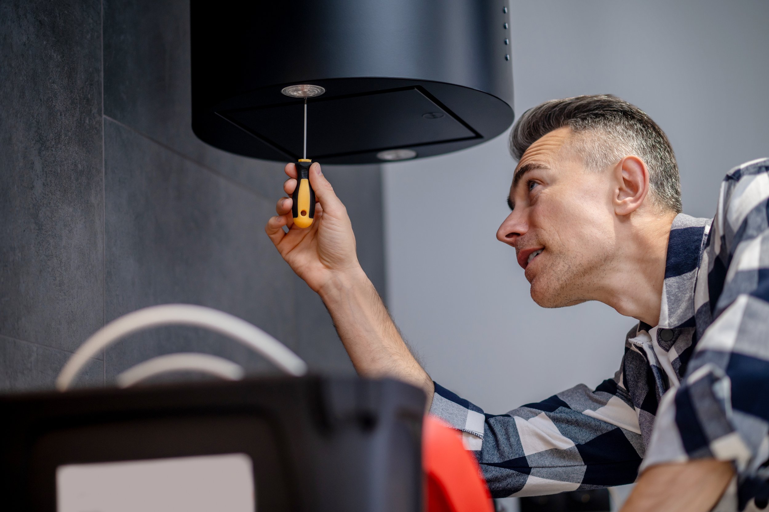 A man wearing a striped shirt is using a screwdriver to work on a black appliance mounted on a grey wall.