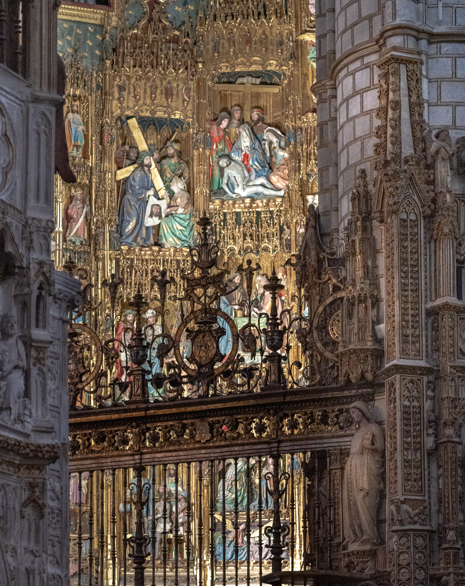 Gilded polychrome altarpiece in Toledo Cathedral seen through a wrought iron grille, its carved scenes lit by warm raking daylight against cool grey stone
