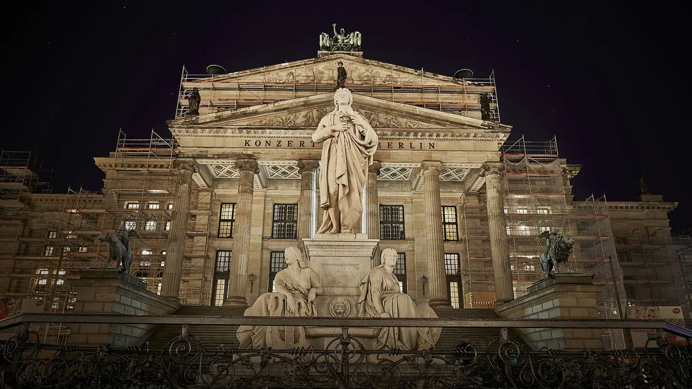 The Schiller Monument and its sculpted figures in front of the illuminated columns and pediment of Konzerthaus Berlin at night.
